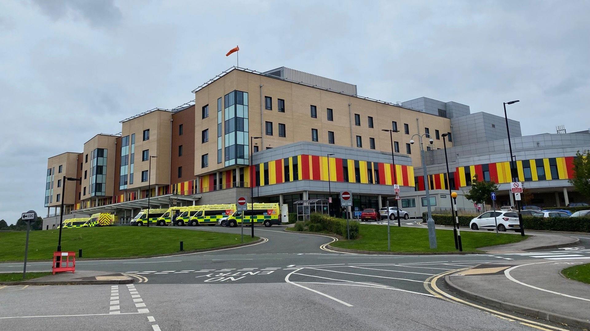 A large light brown-coloured hospital building with a multi-coloured first storey (yellow, red and black stripes). Ambulances can be seen parked outside with a car park to the right.