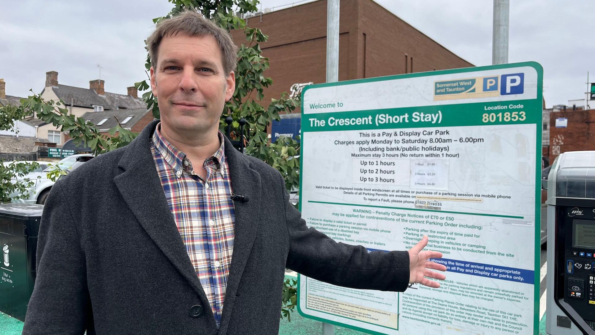 Councillor Wilkins is stood looking at the camera in a car park, gesturing at the car parking board behind him. He's wearing a checed shirt and grey overcoat.