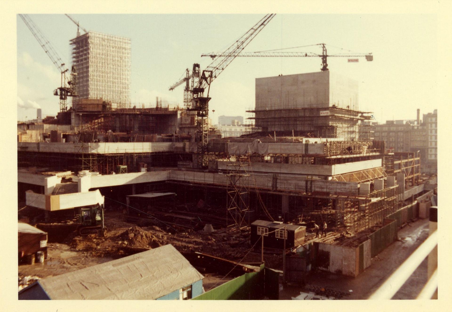 Cranes and scaffolding above the part-built concrete frame of the National Theatre on the South Bank in 1970. There are lots of levels and sections. Behind the construction site are other buildings. 