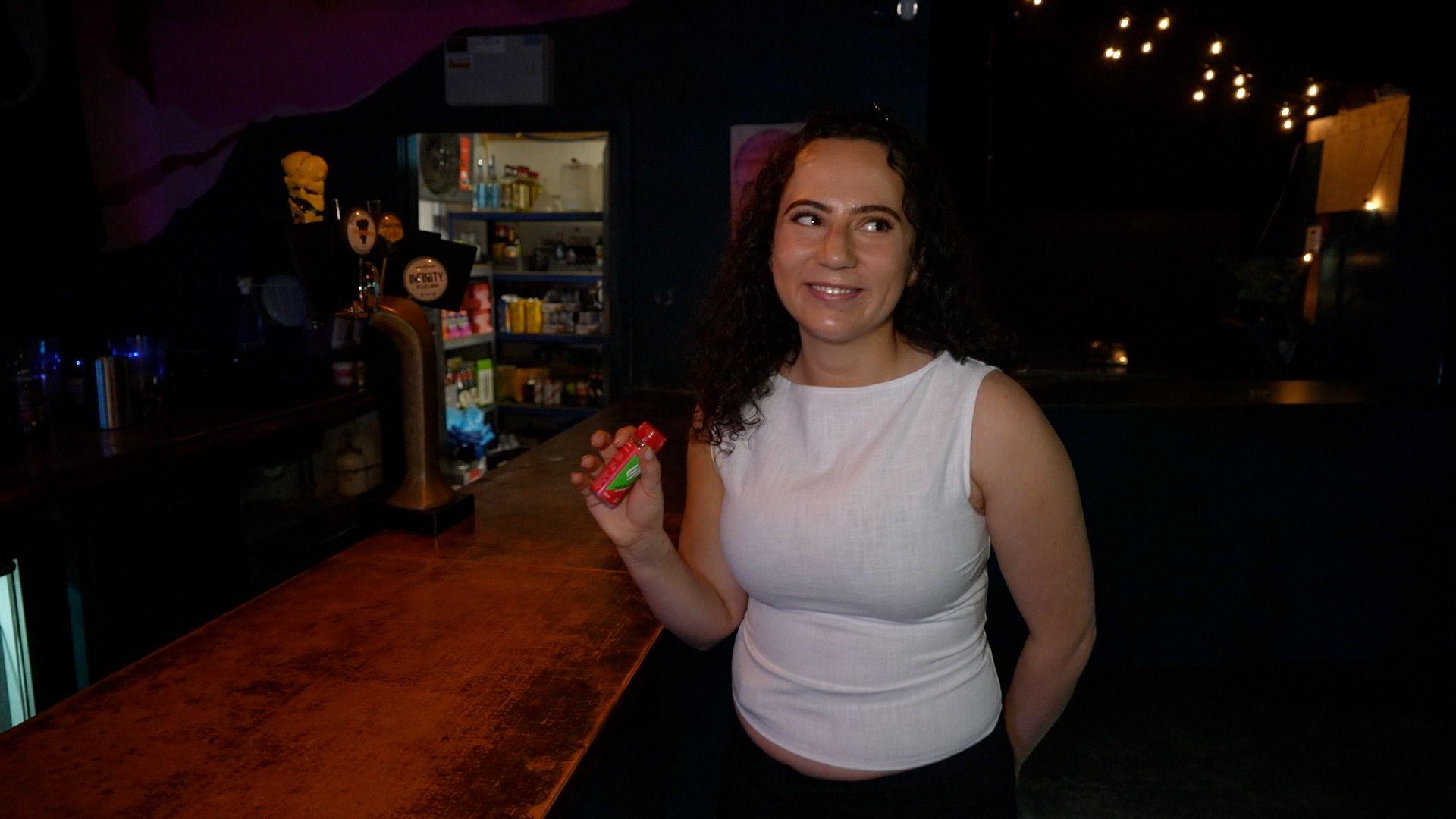 Jessica Unger is holding a Lift glucose shot next to a bar at a club. She wearing a white vest and smiling. The room is quite dark. The beer drafts, pantry and fairy lights in the background brighten up the room a little.