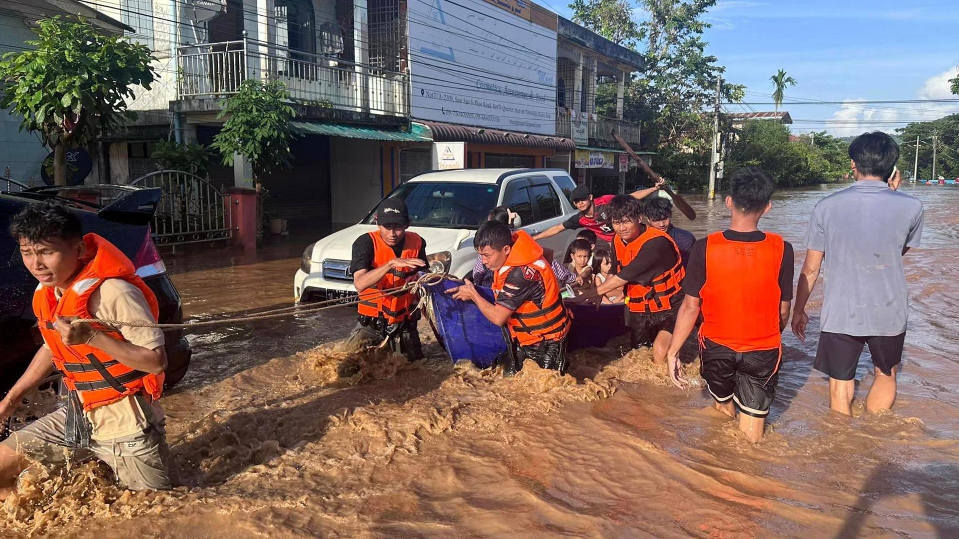 Men in orange life jackets pull along a small boat full of children through waist-high water in a street