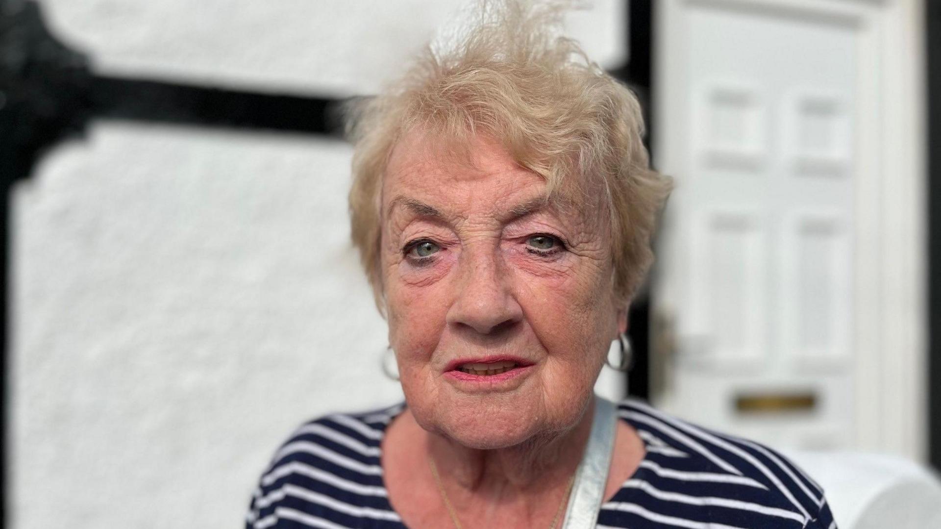 Christine looking at the camera. It is a head and shoulders shot of her. She has wispy blonde hair and green eyes. She has pink lipstick on and you can see the silver shoulder strap of a bag on her left shoulder. She has a blue and white striped top on. Behind her is a blurred image of a white door with a gold letterbox. 