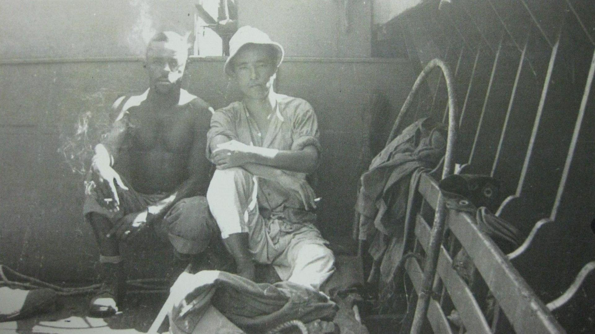 Two men, including Major Yutaka Umezawa, are sitting in the bottom of the boat. Both are smoking. Major Umezawa is in white wearing a white hat, shirt and trousers. The other man, an American soldier, is bare-chested.