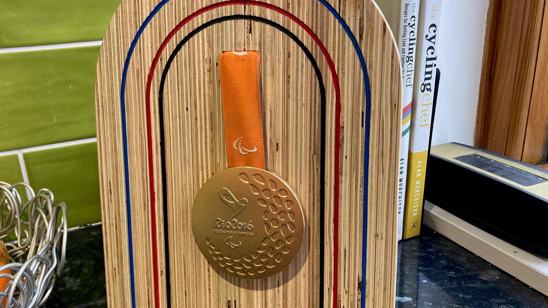 Gold medal for paracycling at the Rio 2016 Paralympics which Steve Bate won in the men's individual pursuit and men's road time trial events at the 2016 Paralympics in Rio de Janeiro with his pilot, Adam Duggleby. The pair also won a bronze medal in the men's road race. The medal is on display on a wooden board on a black kitchen work surface with a metal fruit bowl and green tiles to the left. Cycling recipe books are to the right.