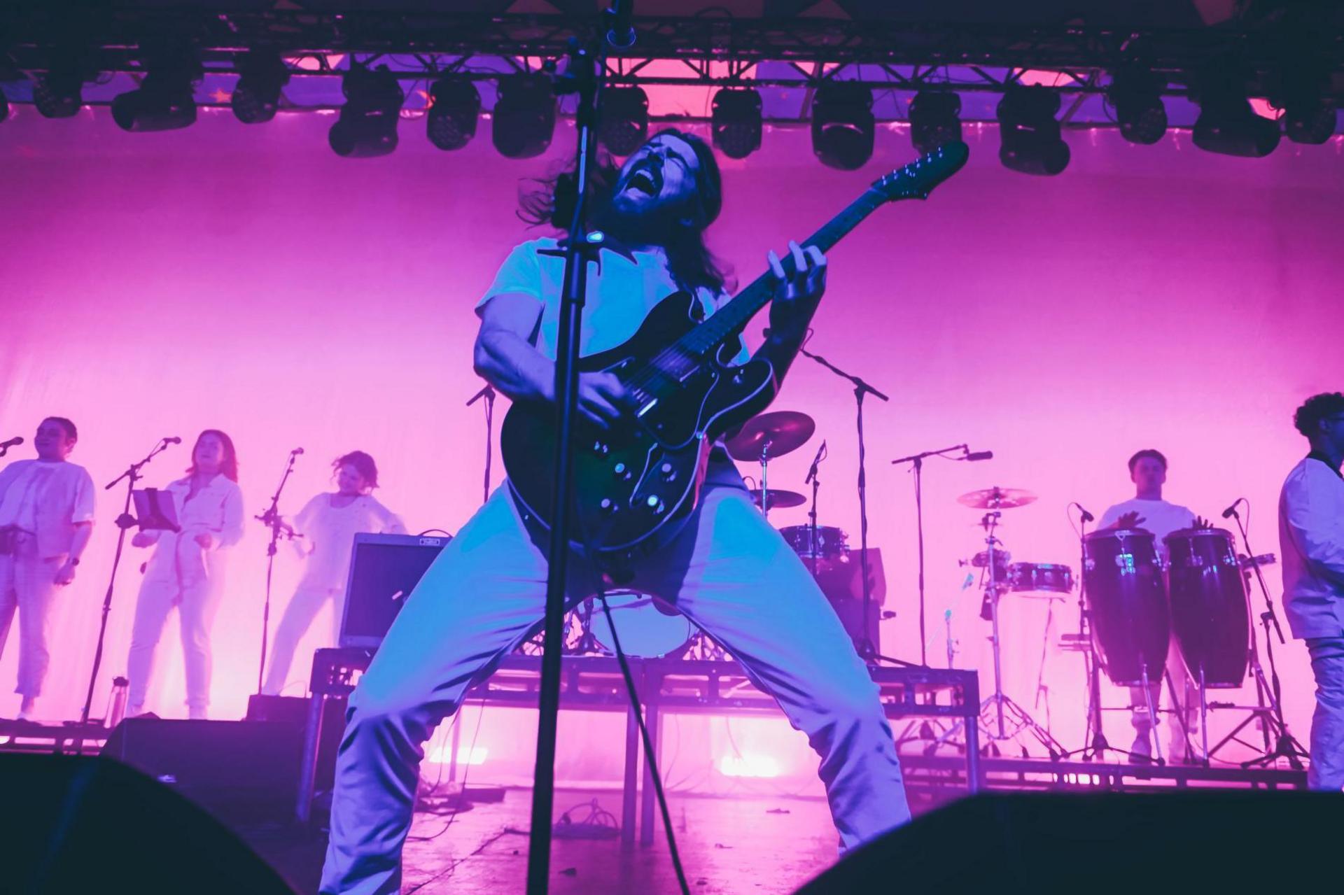 A guitarist and singer in the band screams into a microphone while playing a guitar. Other members of the band are lined up in the background.