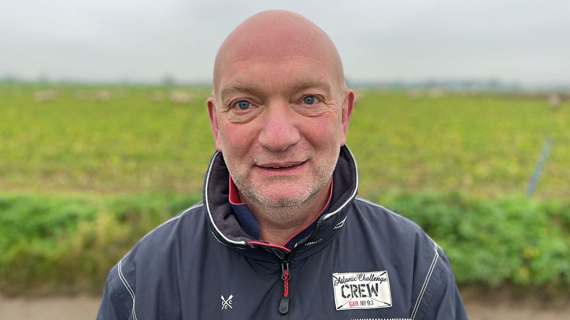 Keith Williamson smiling at the camera while standing outside, in front of a field with sheep. He is bald and has stubble, and is wearing a navy jacket.