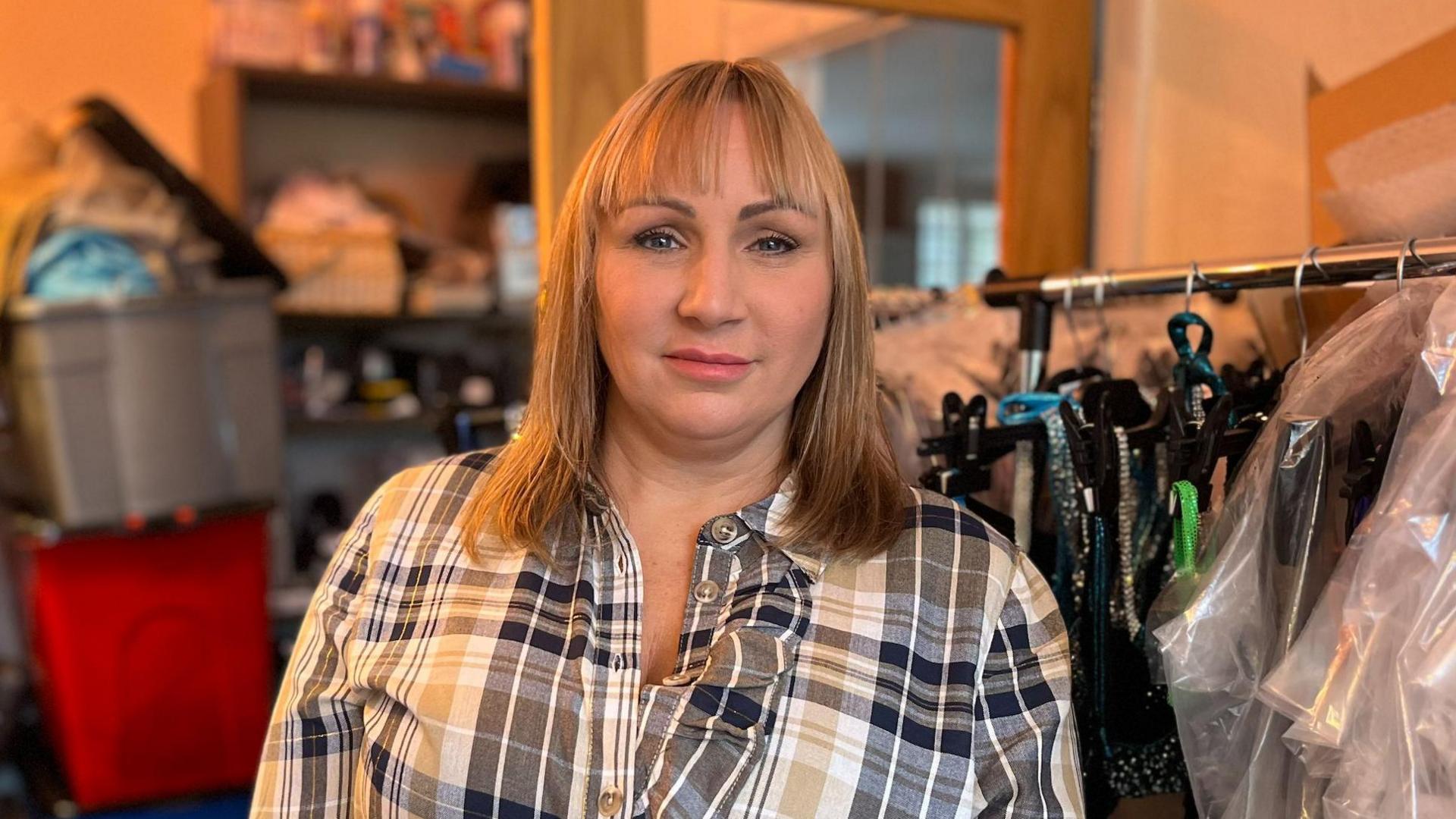 Woman with shoulder length blonde-hair in checked shirt smiling at the camera. She is next to a clothes rail with sparkly outfits.