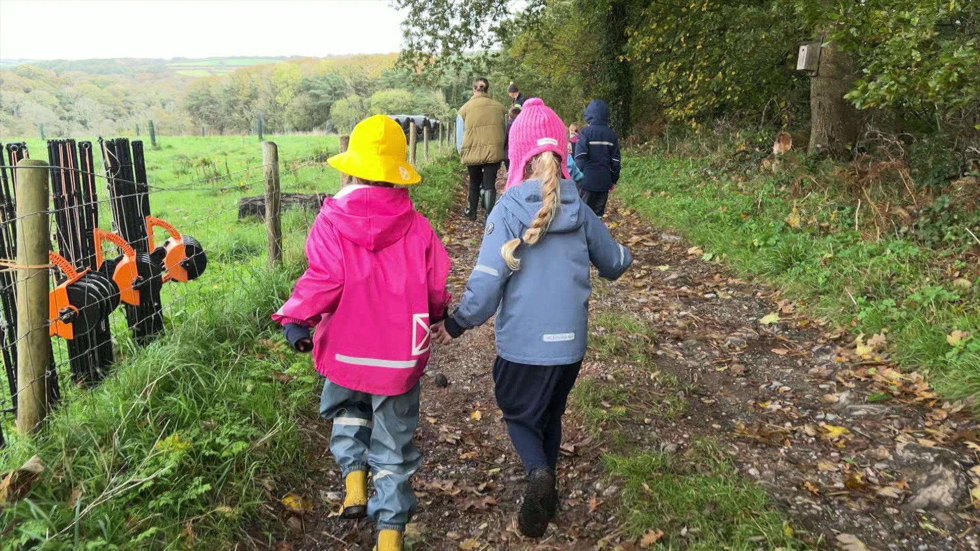 The back of two children walking and holding hands. They are both wearing hats and coats as they walk down the pathway in a field. To the left are wooden posts with metal fencing between them. On the right is grass and trees.