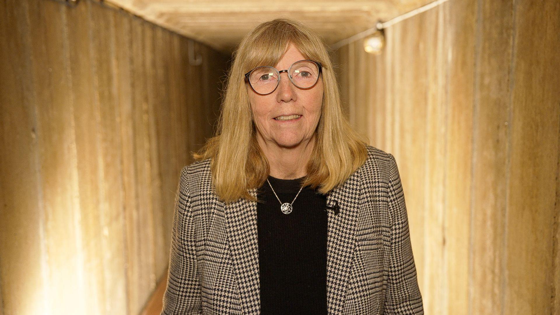 A woman with shoulder-length blonde hair. She is wearing a black shirt with a black and white blazer on top. She is standing in the underground tunnel.