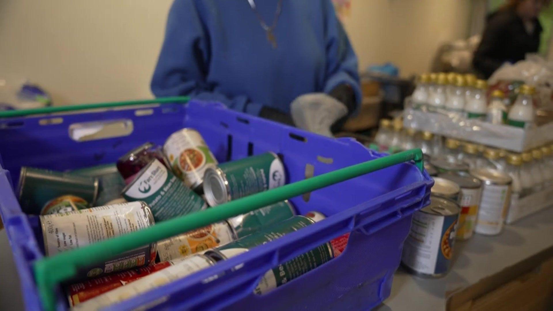 A close-up image of a box full of tinned food, with someone in the background packing tins into a bag