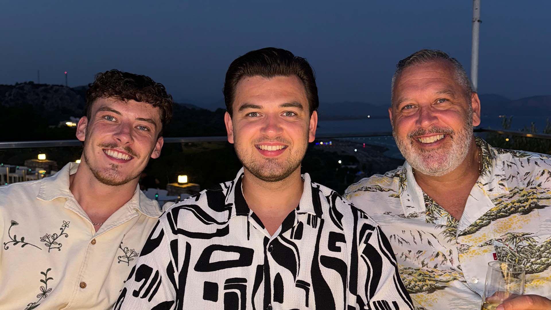 Travis, Harrison and Paul, all smiling, looking at the camera. Travis has a cream and black patterned shirt on, Harrison has a black and white shirt on and Paul, has a floral shirt on and is holding a glass.