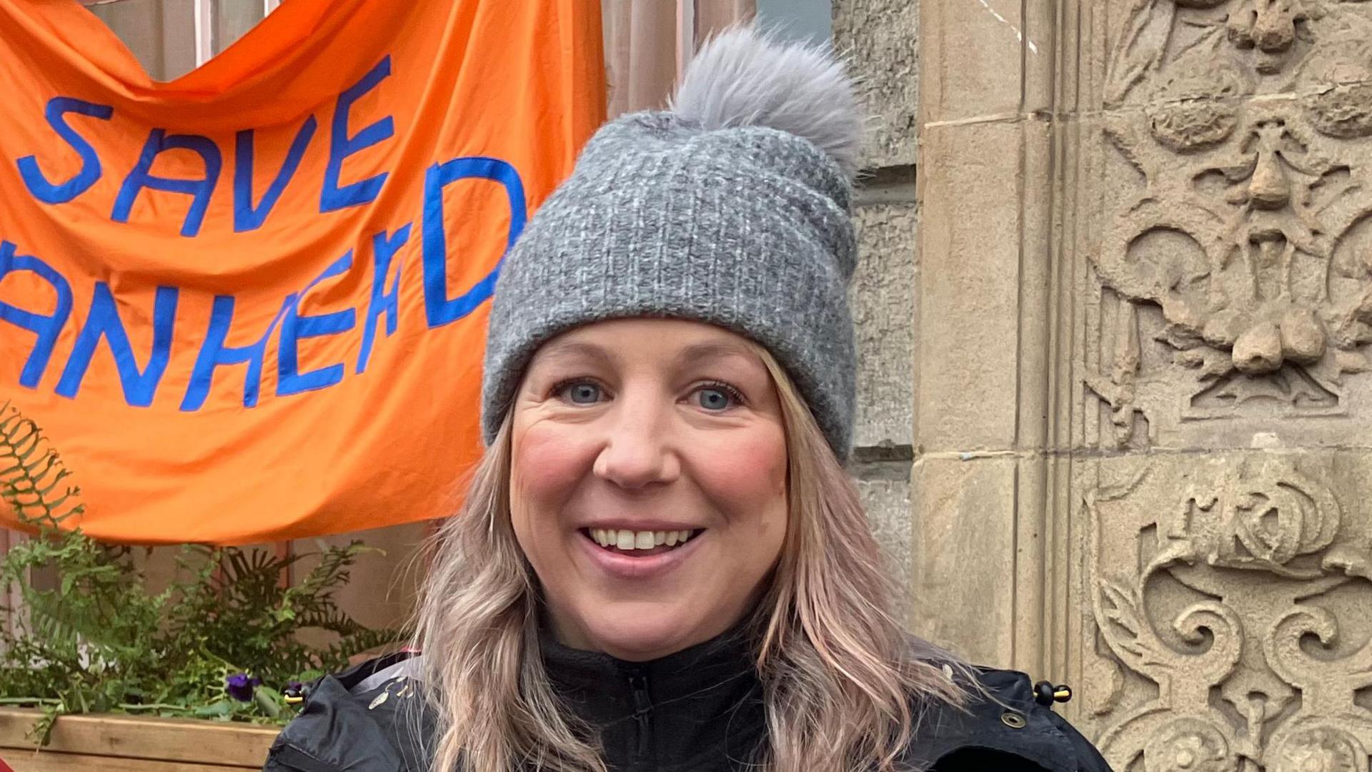 Leanne Parr, who has long blonde hair and is wearing a grey bobble hat and a black coat, is standing outside a stone building. An orange banner with blue lettering can be seen behind her, reading 'Save Roanhead'. 