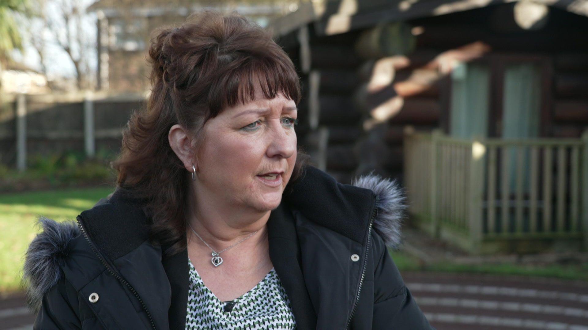 A woman with reddish brown hair looks to the right of the camera, wearing a coat and standing in the hospice gardens 