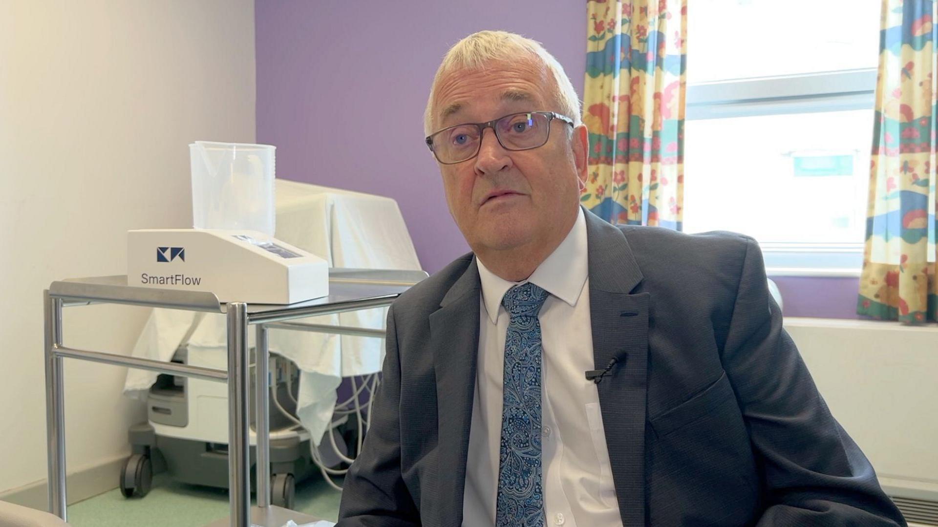 Cyril Barrett, wearing a grey suit jacket, white shirt and blue patterned tie. He is sitting in what appears to be a hospital room