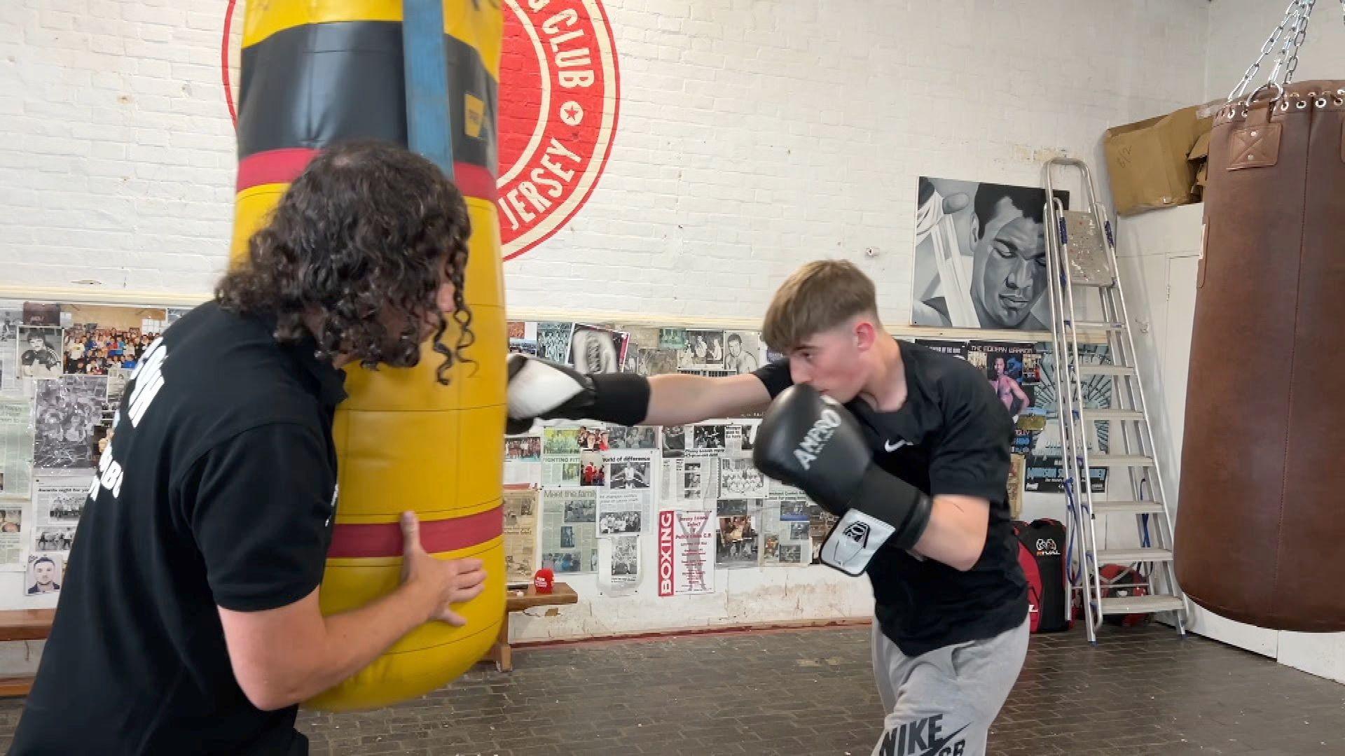 A teenage boy punches a heavy boxing bag that's hanging from the ceiling while a coach holds the bag in place