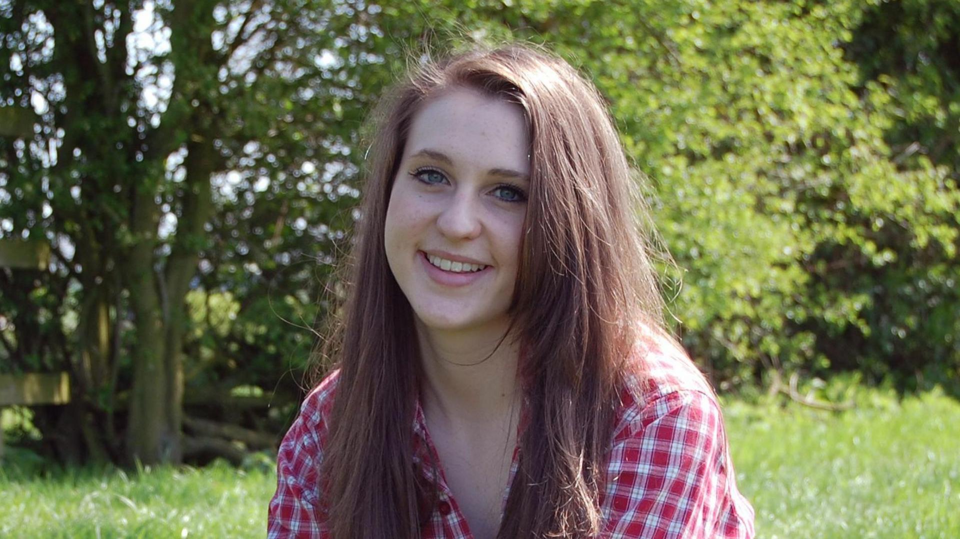 Alice Ruggles smiles at the camera. She has long brown hair and blue eyes and is wearing a red and white checkered dress. She is sat in a field of long green grass with trees and bushes in the background.