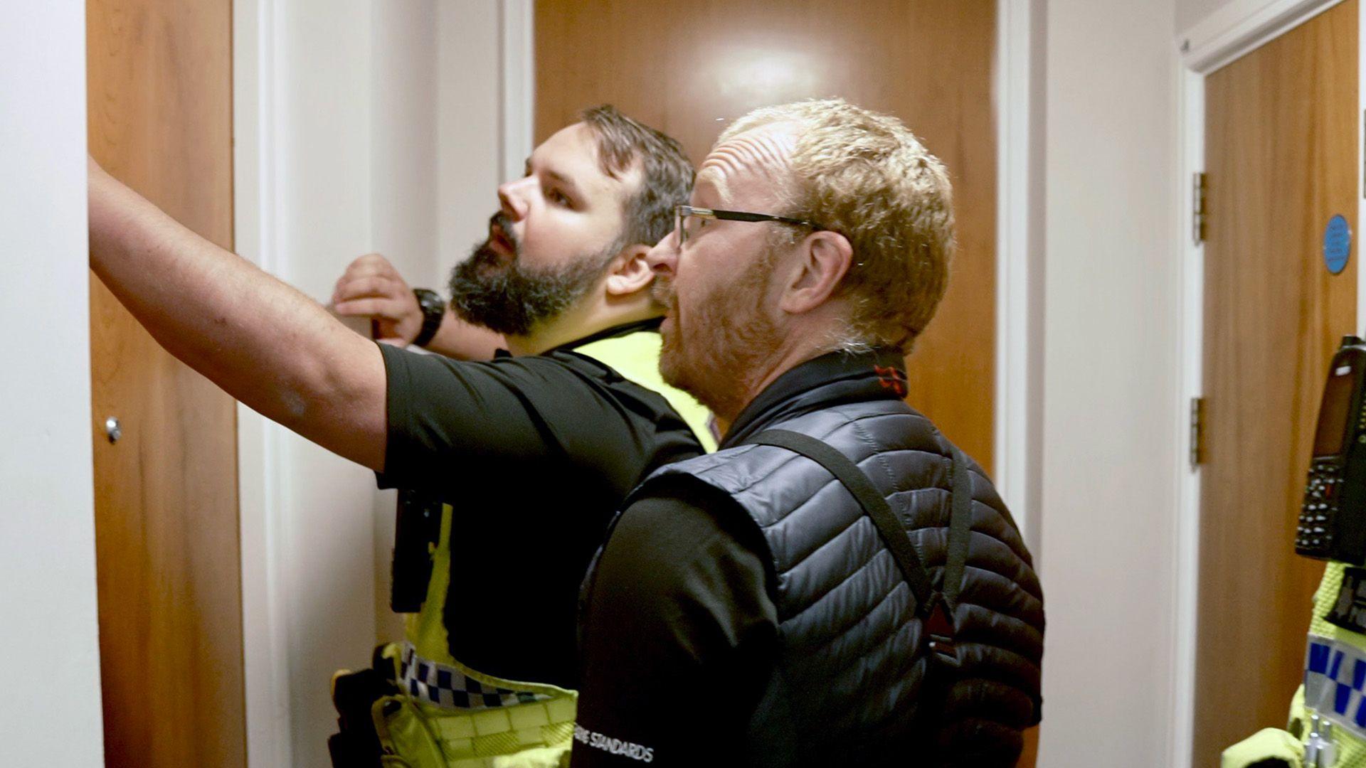 Rhys Harries stands in a hallway wearing a black quilted jacket labelled "Trading Standards". In front of him a police officer puts his hand on the door.
