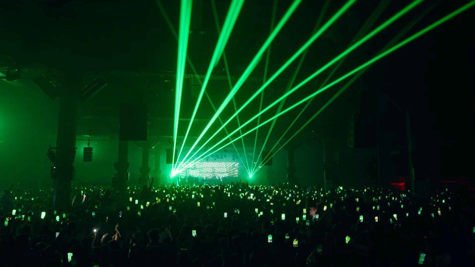 A huge crowd at a gig at The Warehouse Project, Green lights shoot across the ceiling, and a sea of phones can be seen in the crowd as people record the DJ.