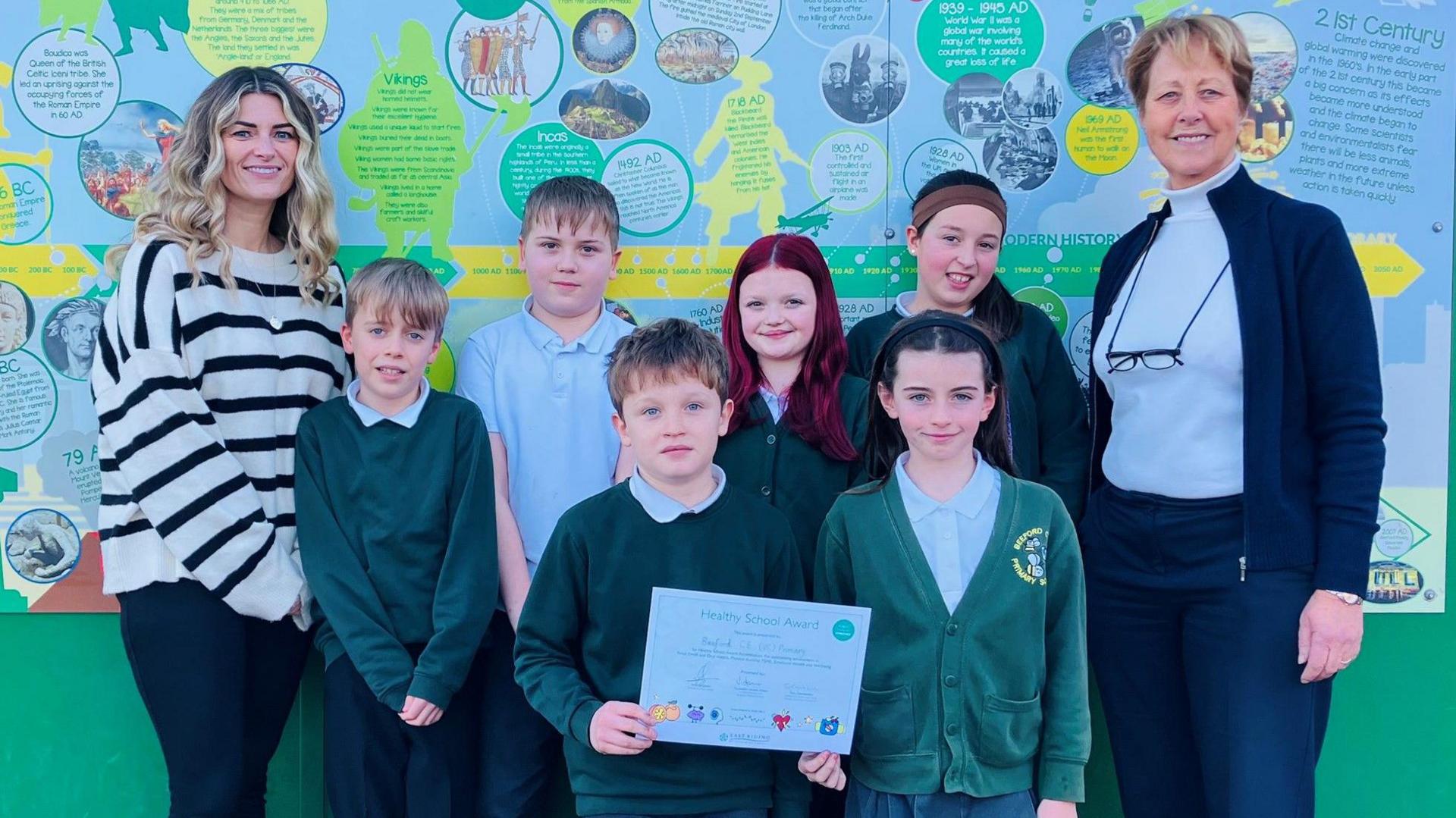 Six children hold a certificate to celebrate their school being given a health award. Mel Wilson, the school safeguarding lead, stands on the left of the image - she is wearing a blue and white striped jumper and has wavy blonde hair. Councillor Victoria Aitken is standing on the right of the image. 