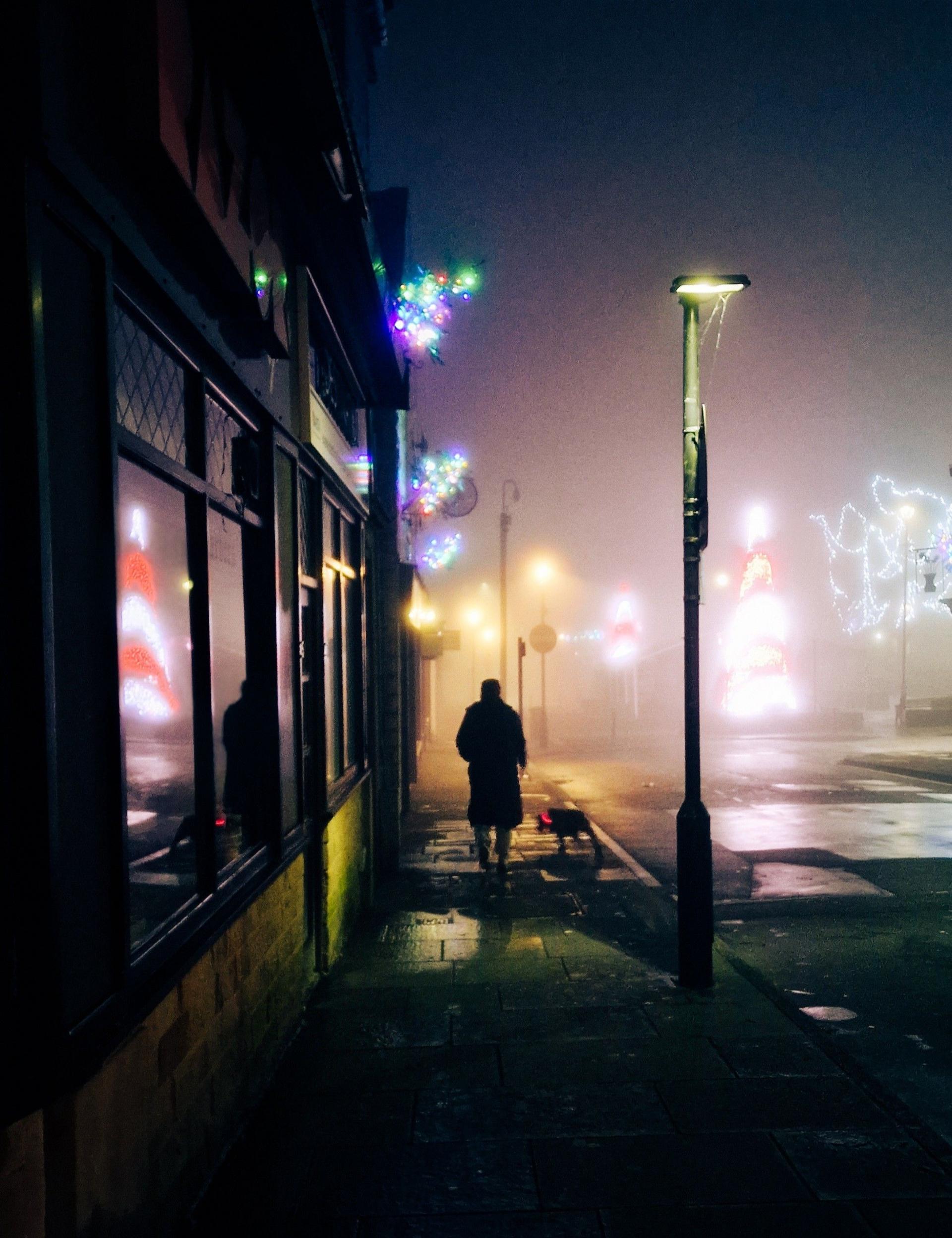 A figure walks along a foggy road in the dark. There are buildings on the left of the figure, a road on the right and Christmas lights in the distance.