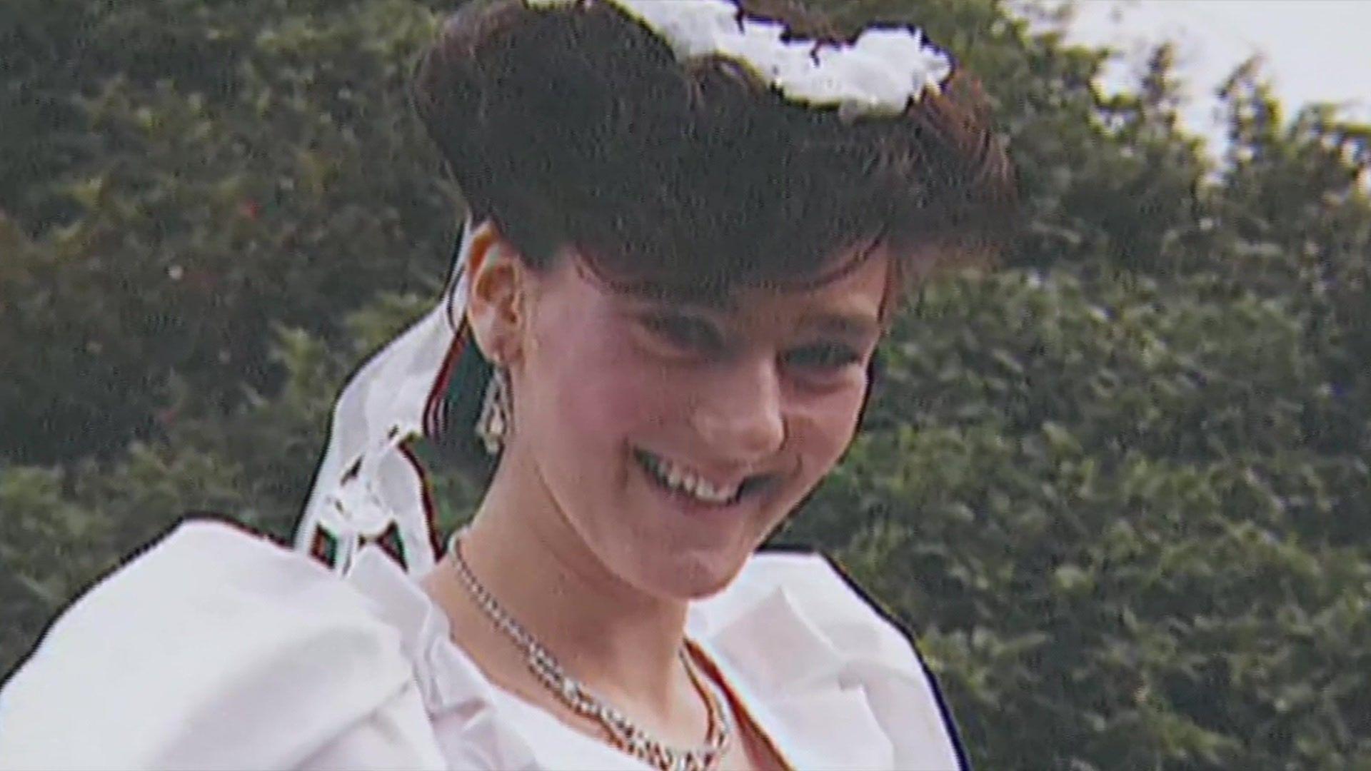 Julie Hogg smiles at the camera. She is wearing a fancy white dress and white crown atop her short thick dark hair.