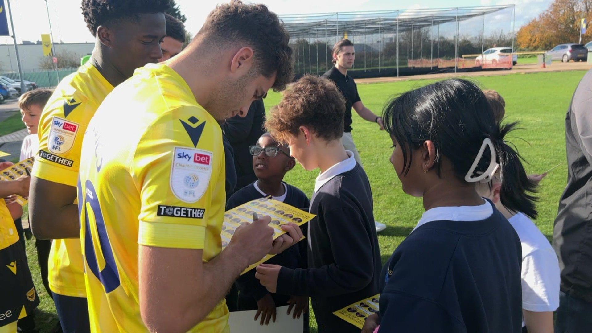 Two Oxford United players wearing yellow t-shirts sign sheets of paper for a group of Year 6 students.