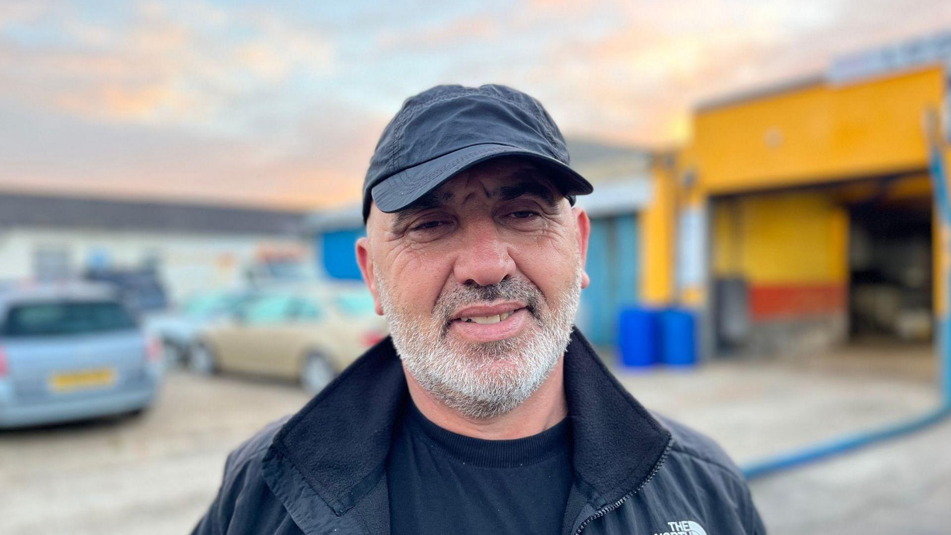 A middle-aged man with short grey stubble. He is stood in the car park outside a yellow car wash garage and is wearing a black t-shirt, black cap and black jacket