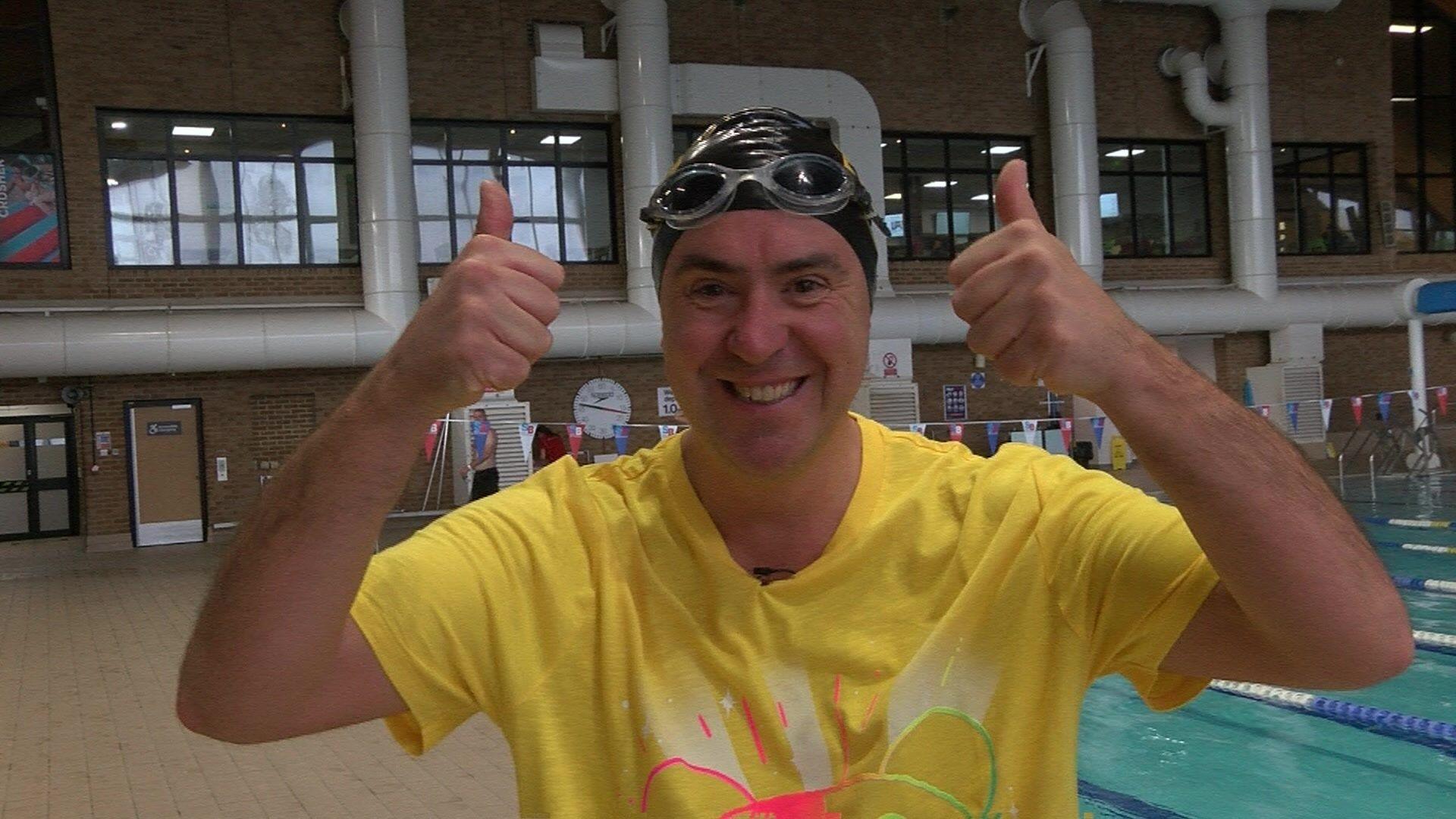 A man in a yellow t-shirt, a black swimming hat and goggles standing in front of an indoor swimming pool, smiling and giving a thumbs up