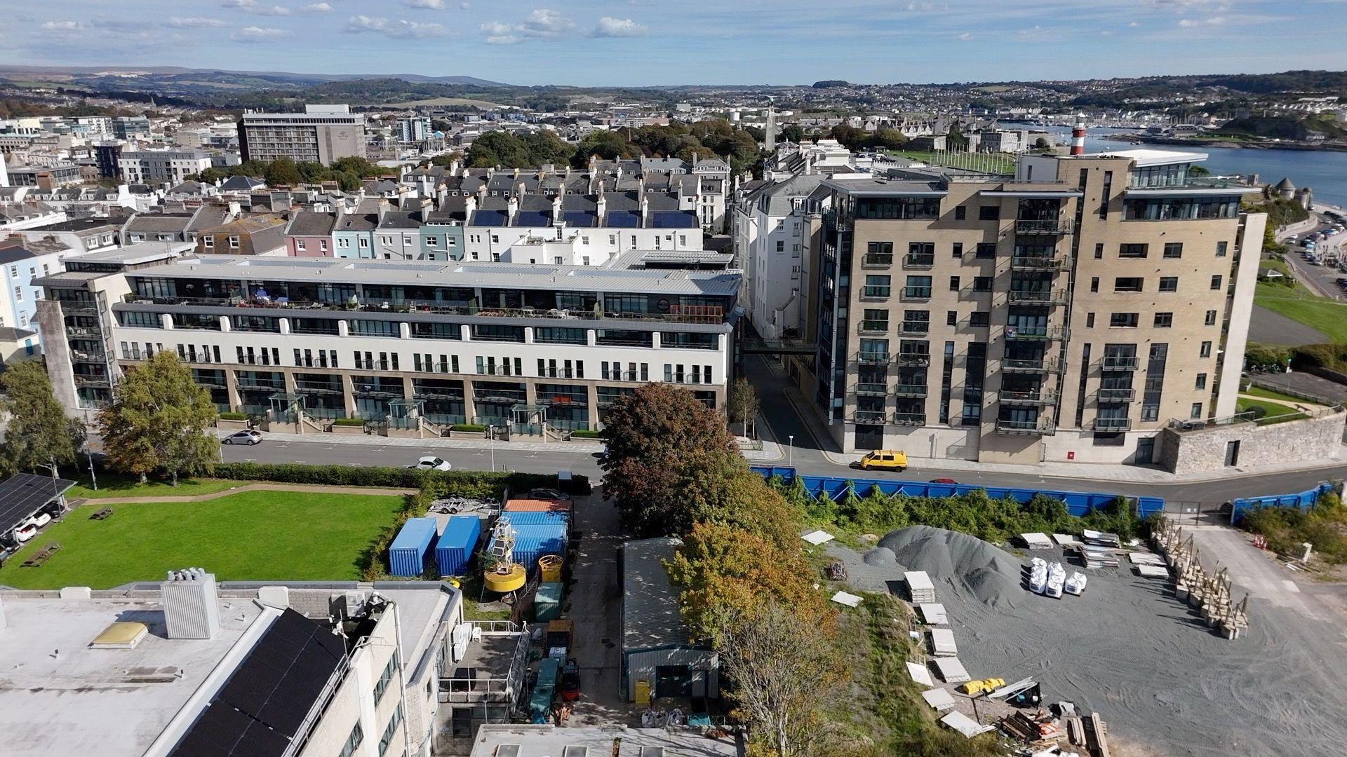 A wide view from a drone of two buildings with a walkway between them barely visible in the shadows. The building on the left is long and low at about five floors, while the building on the right is more square-shaped and has seven floors. The roof of a building, a builder's yard and a patch of grass are visible in the foreground. The sky is blue and housing and other buildings can be seen in the background as well as - to the right of the frame - part of Smeaton's Tower, the red and white lighthouse on Plymouth Hoe, and a sliver of the blue waters of Plymouth Sound.