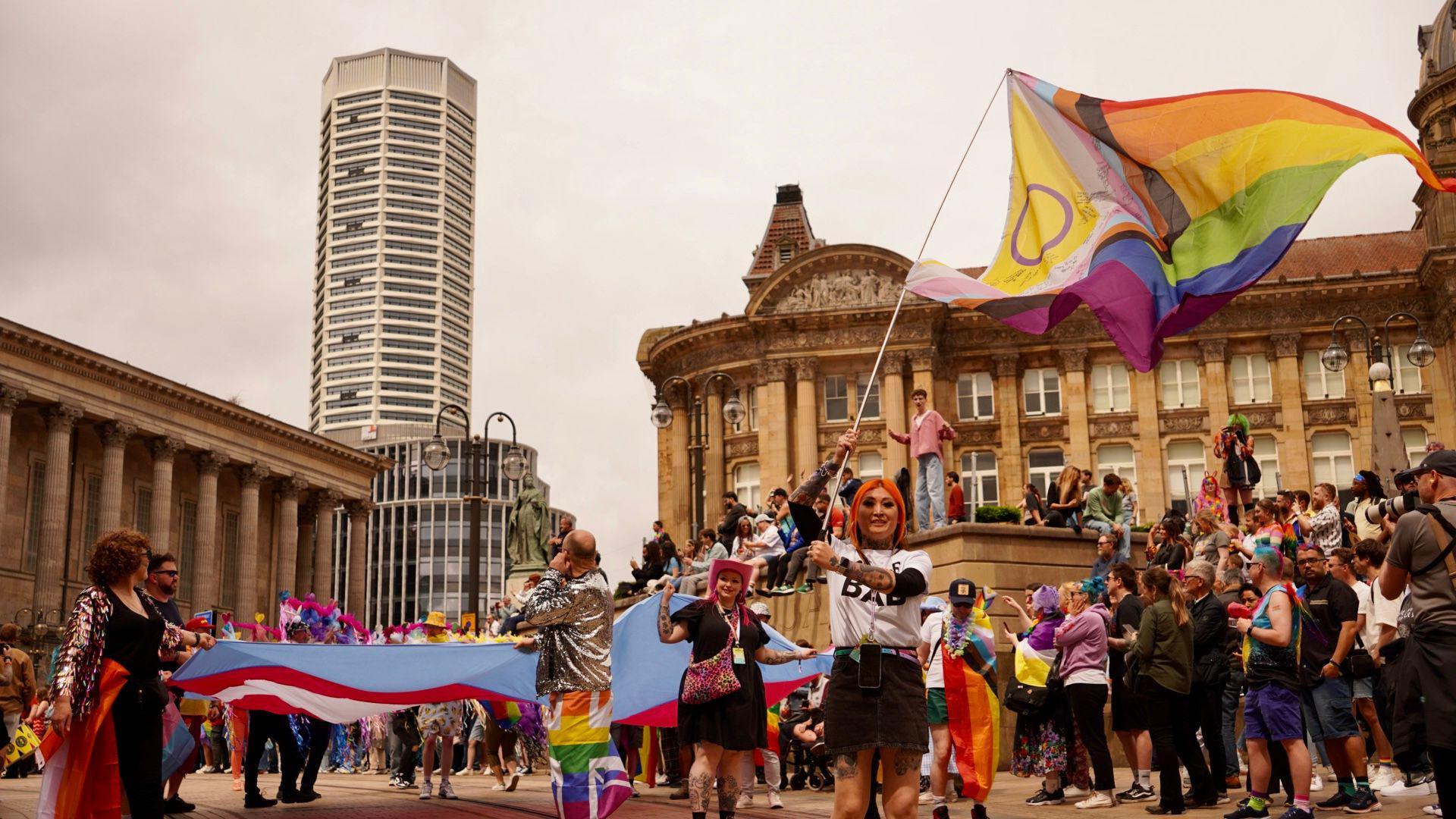 A woman with orange hair waves a rainbow flag in a wide shot Chamberlain Square Birmingham. She is wearing a white t-shirt and black mini skirt. Behind her are crowds of people wearing rainbow accessories. Birmingham's council house can be seen to the left and the Octagon tower behind.