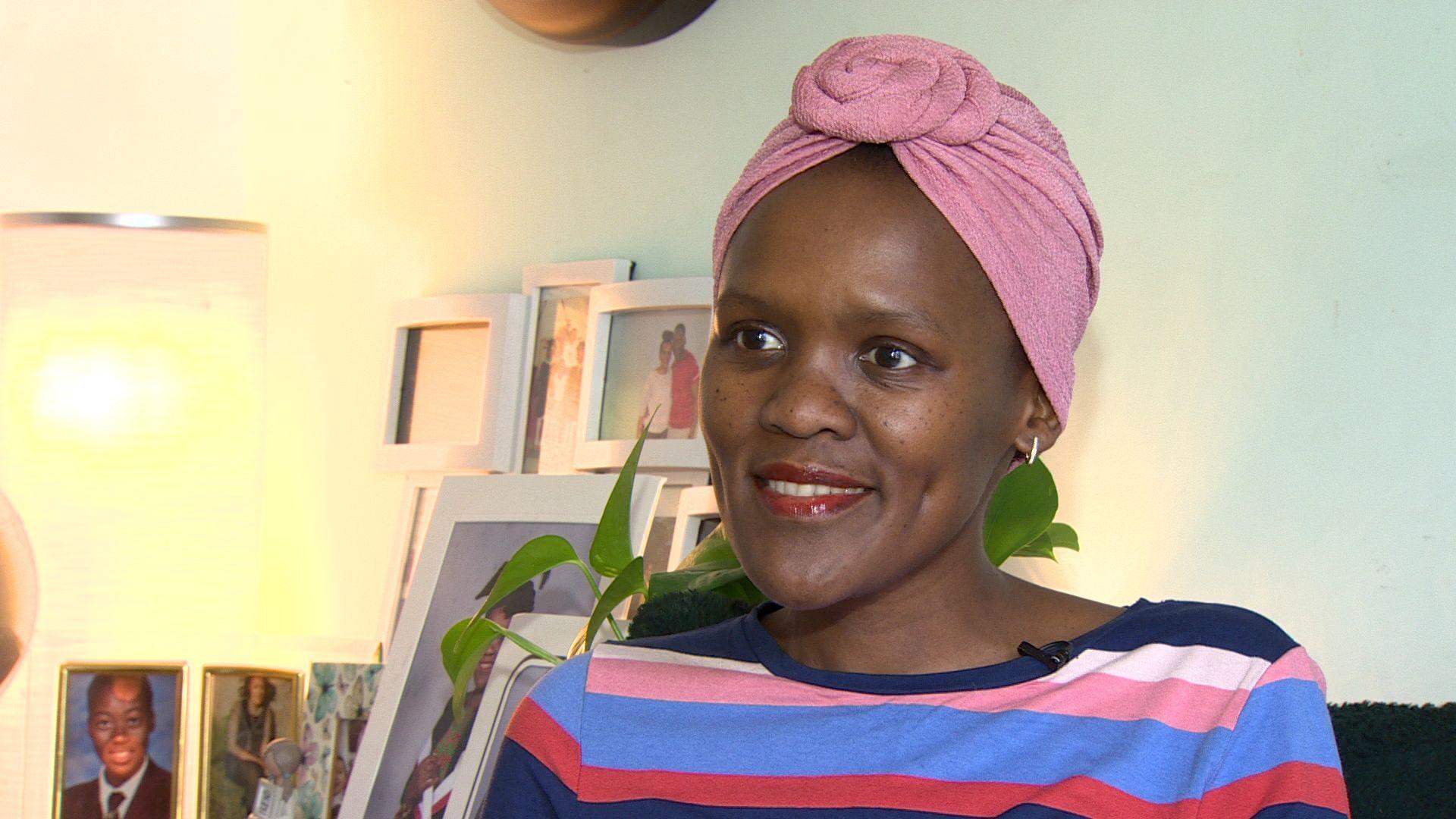 Amanda Suka, wearing a pink headwrap and a striped top, with photographs in the background