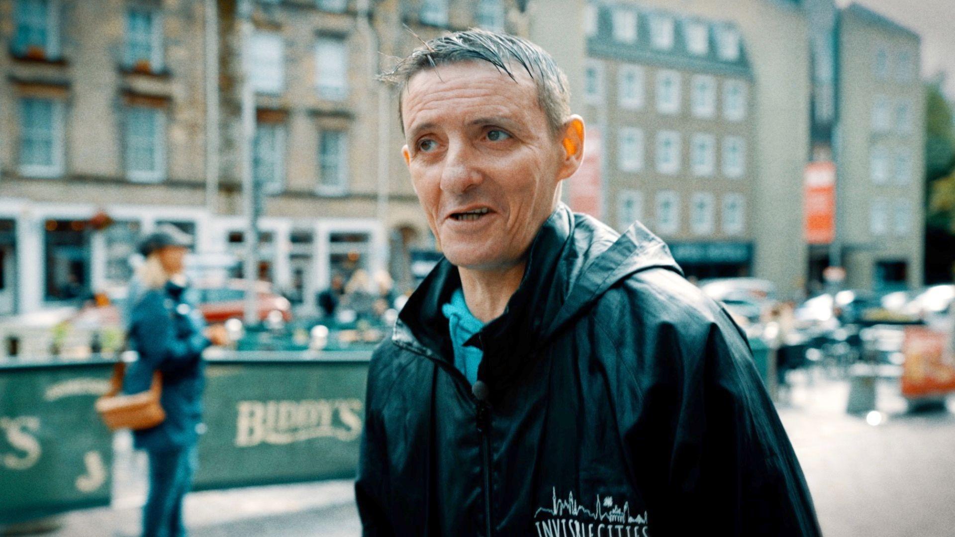 Sonny, who has short black hair and is wearing a black rain coat, walking through Edinburgh's Grassmarket area during one of his guided tours of the city.