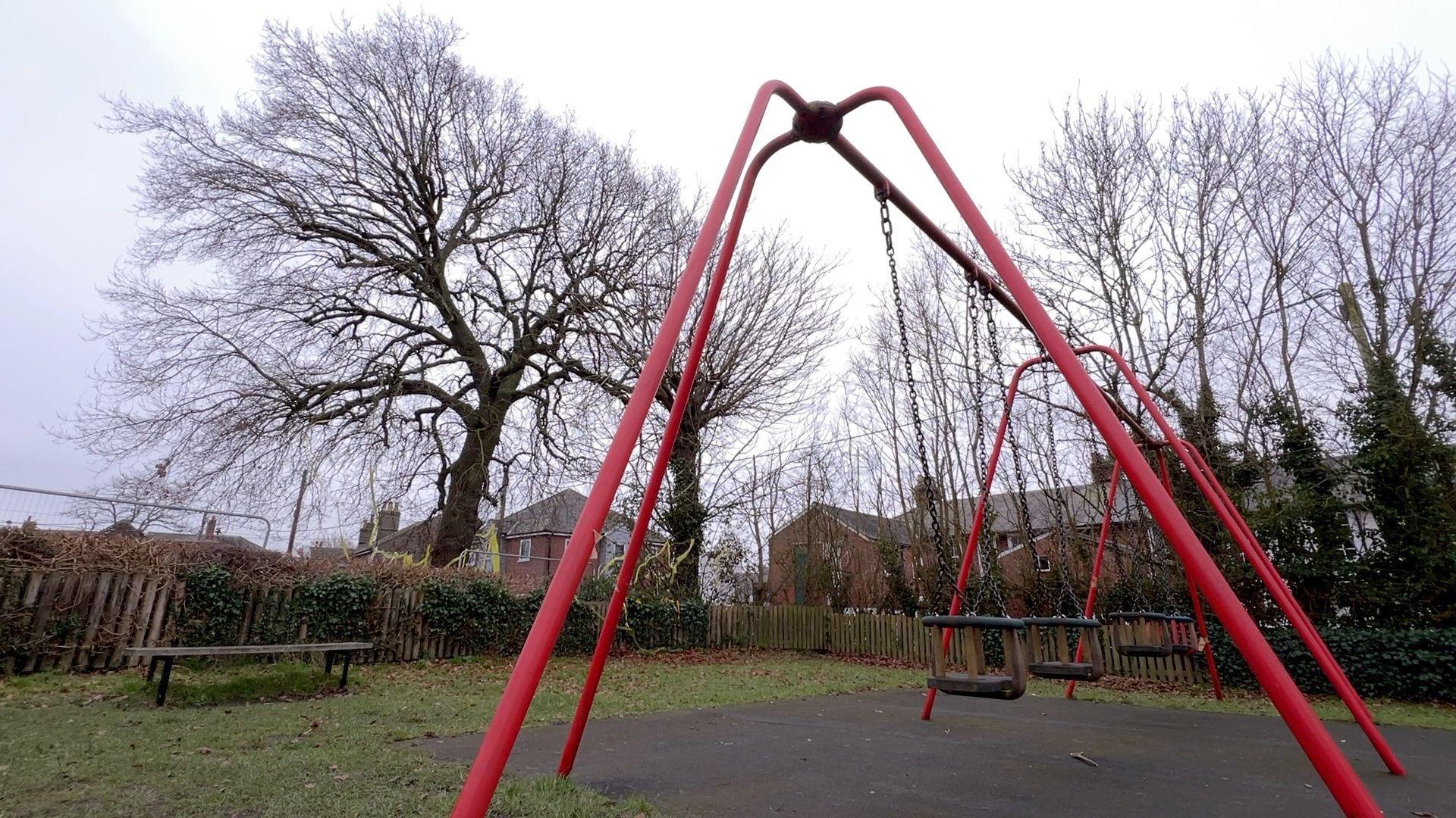 An oak tree, pictured next to a playing field. There is a set of red swings in the foreground and a bench to the left-hand side.