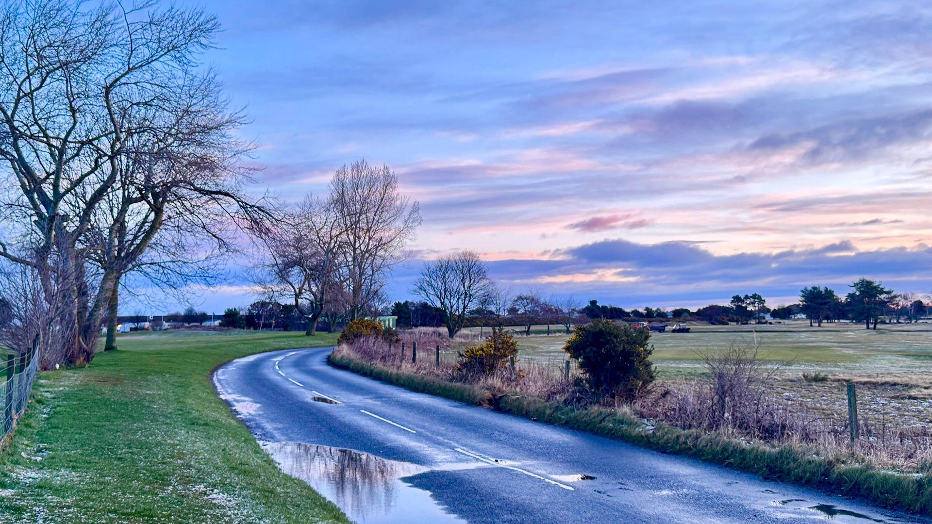 A country scene in Nairn in the highlands with fields and a country road winding away in to the distance. There is a pale sky with pinks and greys. No people or buildings are visible