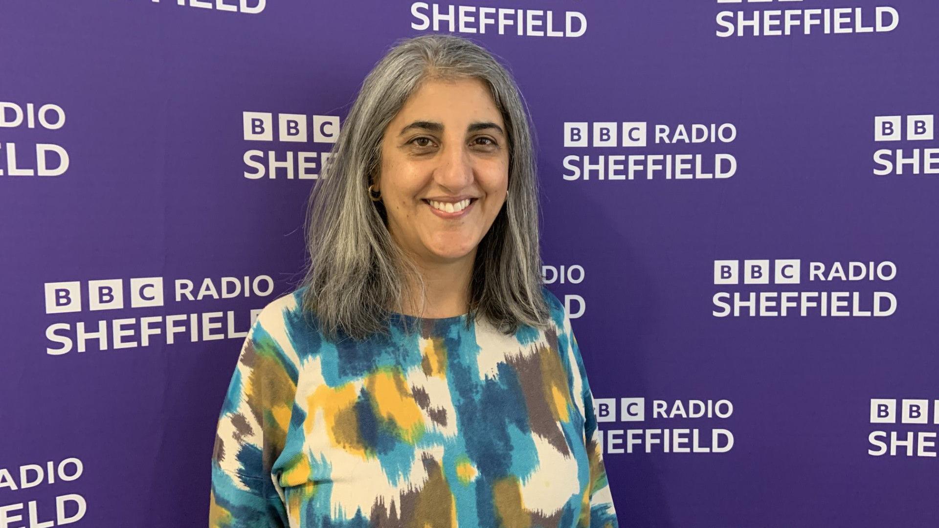 A woman with shoulder length grey hair is wearing a blue, yellow and white patterned top. She is standing in front of a purple wall with white writing saying BBC Radio Sheffield