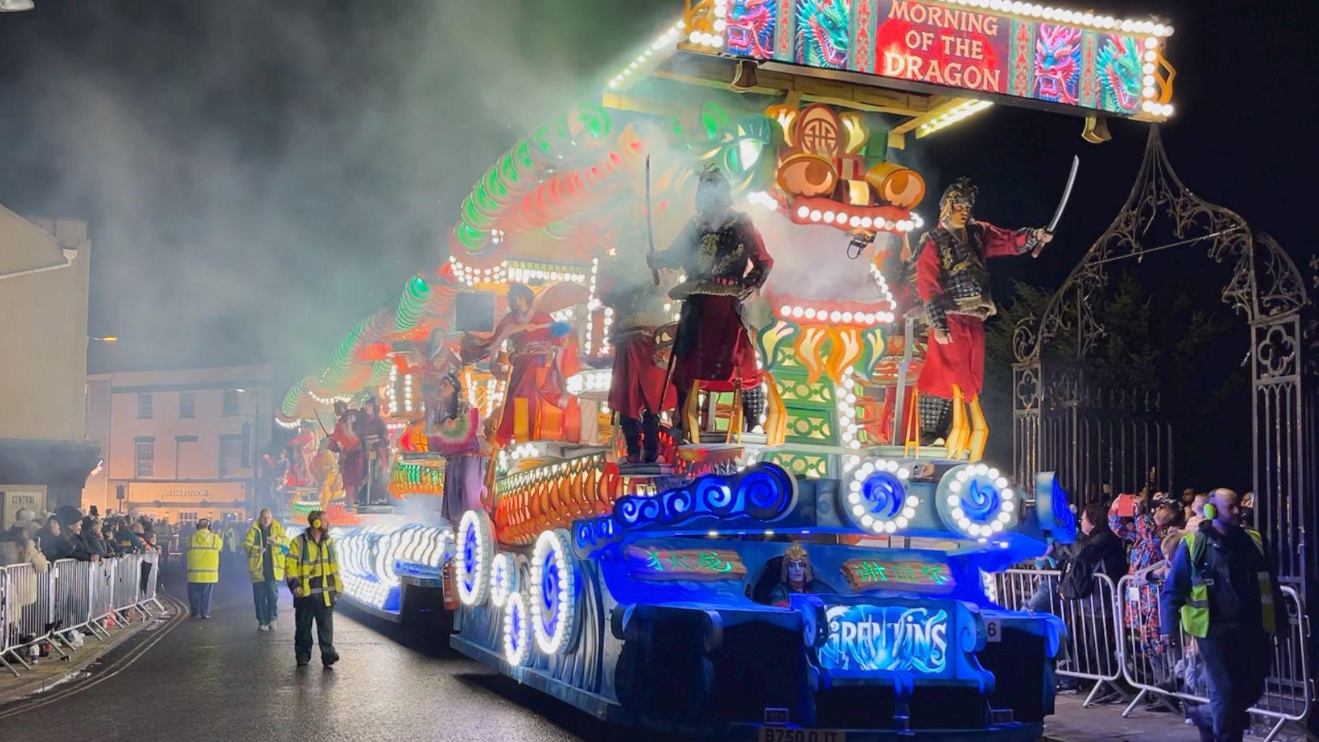 A Gremlins Carnival Club float is being driven down a road on the night of the Bridgwater Carnival. The float is decorated with several bright colourful lights, most notably blue, green and red. There are several performers standing on the float. Spectators are watching from behind metal barricades on the street