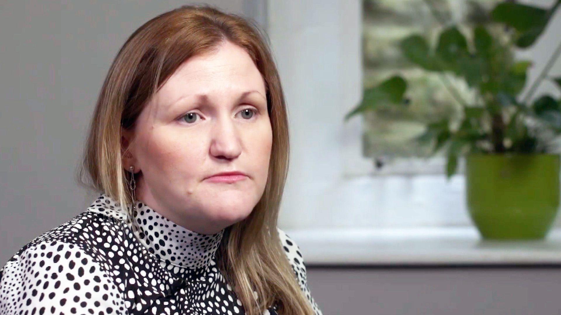 A photo of Karen Booth, looking serious and deep in thought. Her blondish-brown hair is loose and she is wearing a spotted black and white top. A pot plant is in the background on a white windowsill. 