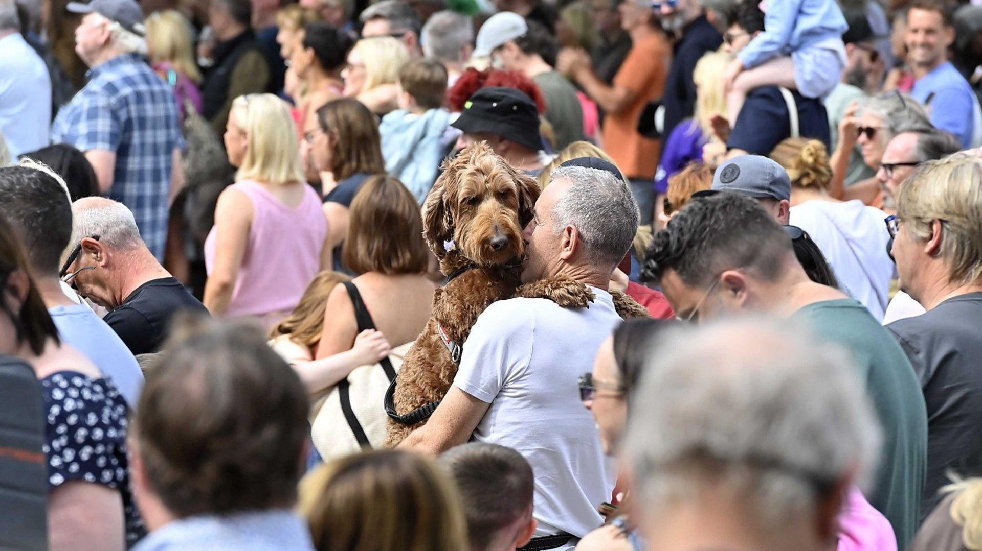 A man is holding a dog. The dog's paws are over his shoulders. They're among a large crowd.