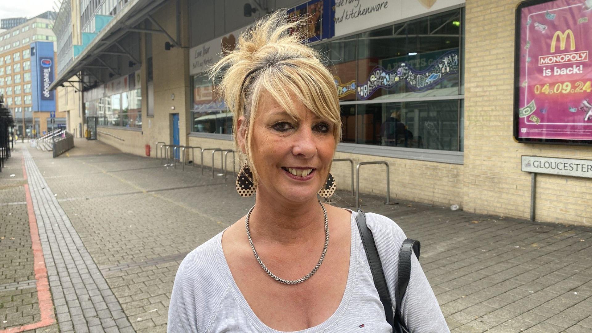 Lorraine Wooton outside the Bull Ring Indoor Market. She is smiling and wearing a low cut top with a necklace. 