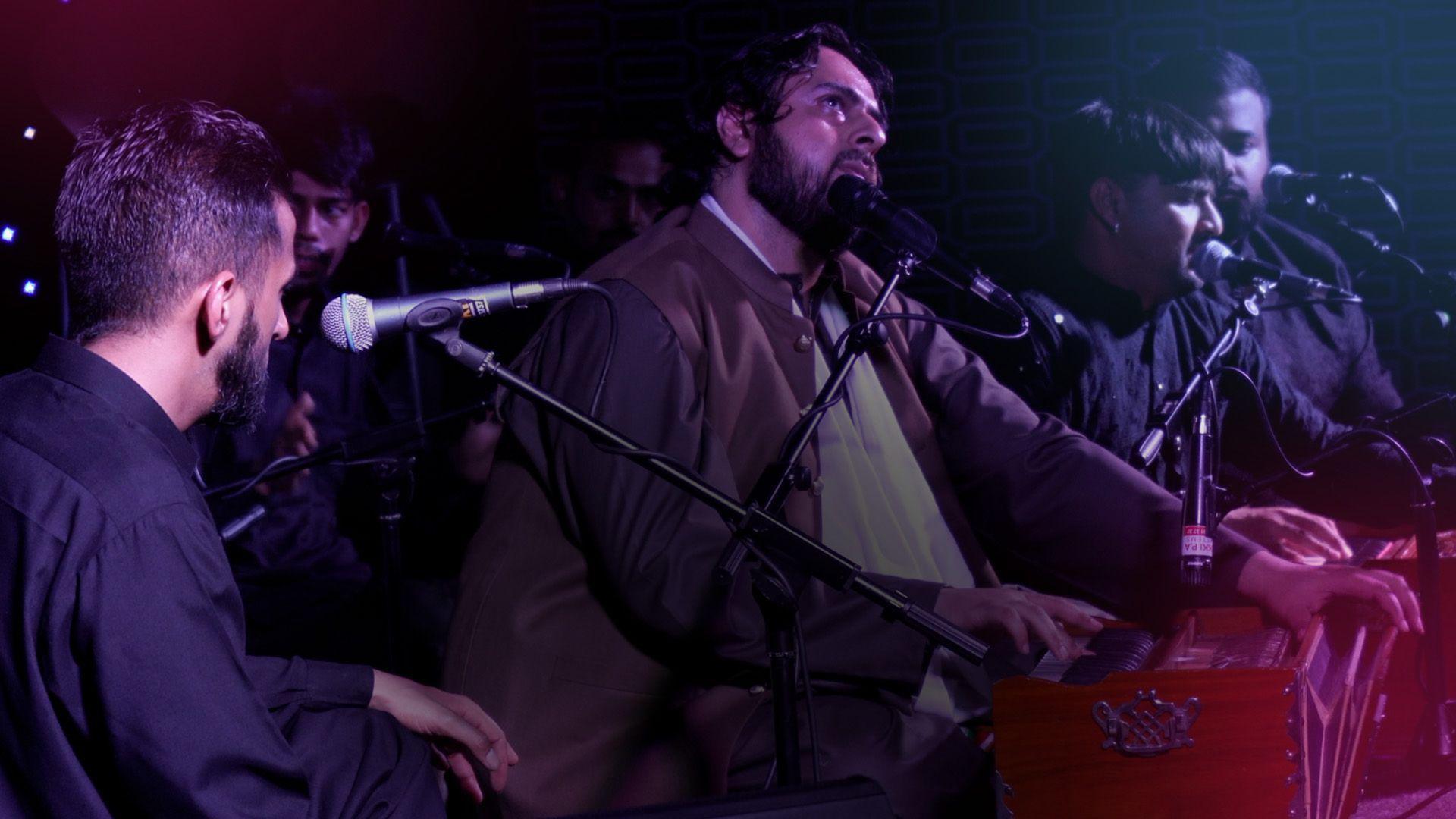 A group of men sitting on a stage with microphones on stands in front of them. The man in the centre is playing the harmonium. All the men are dressed in traditional South Asian clothes.