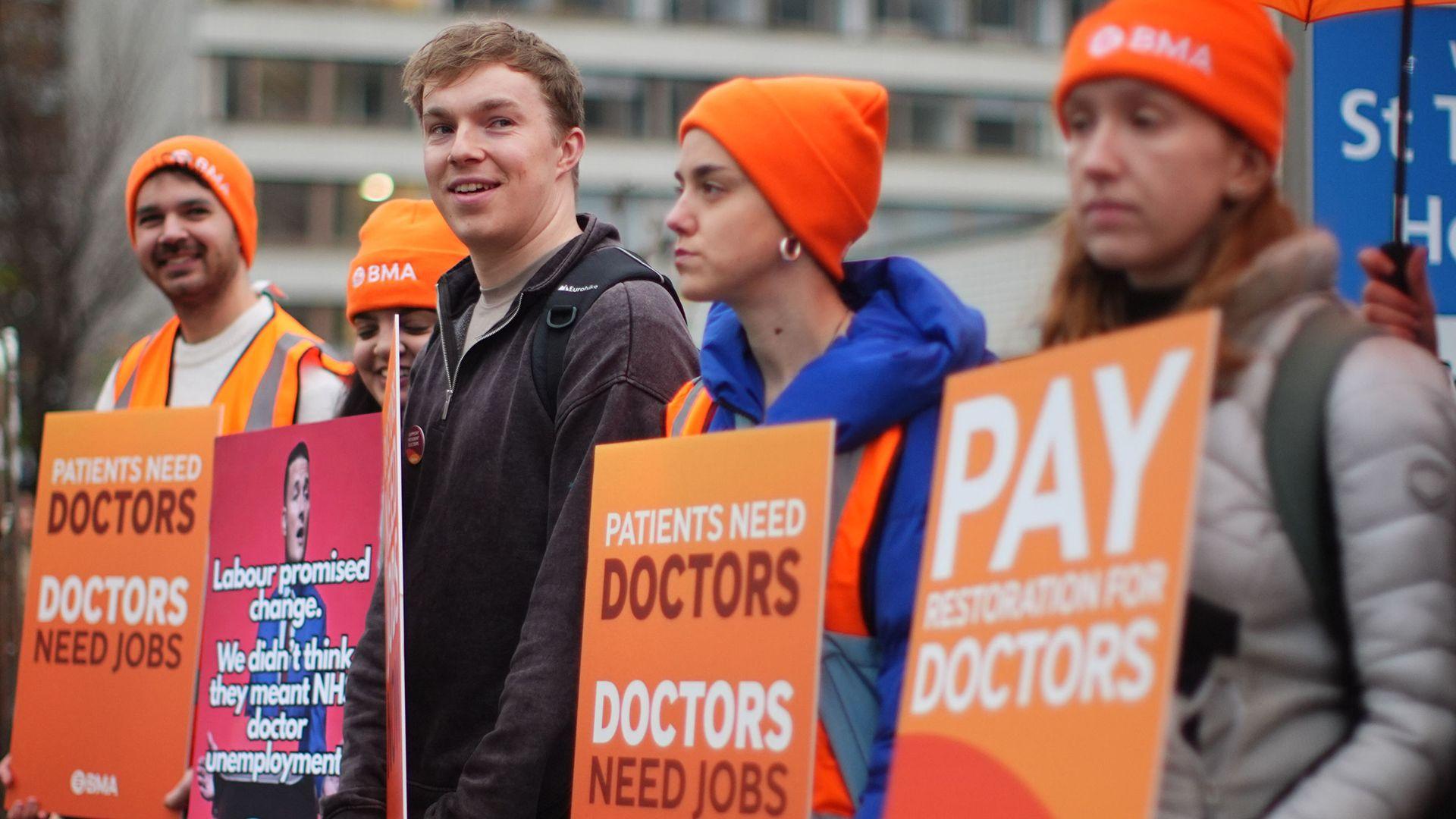 Several doctors on strike, wearing orange BMA hats and holding up placards calling for more pay, saying 'patients need doctors, doctors need jobs''. In the background St Thomas' Hospital is visible. Taken in London on Friday.