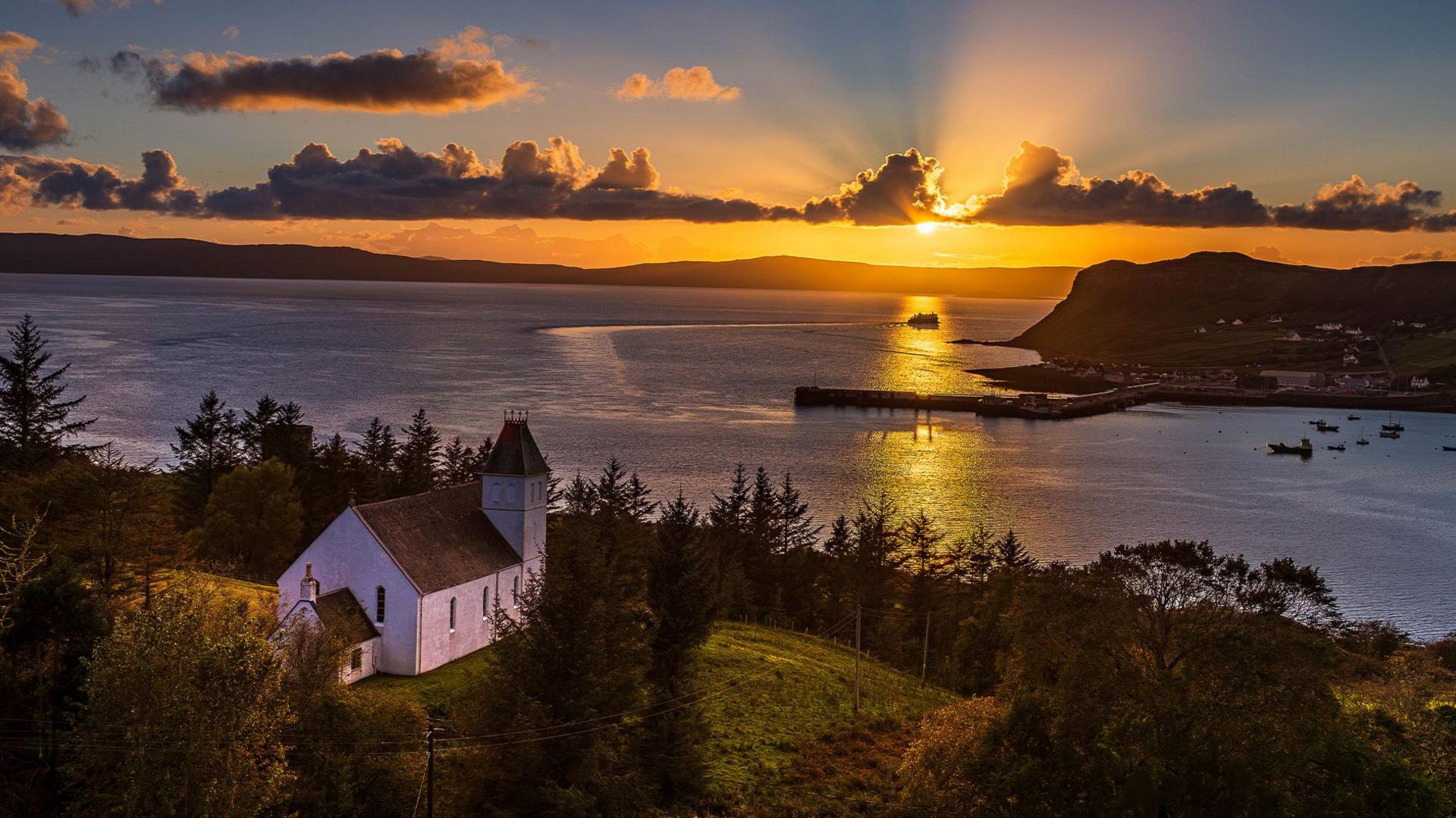 Sun setting over a tranquil bay, surrounded by trees with a white building perched on the hillside overlooking the water.