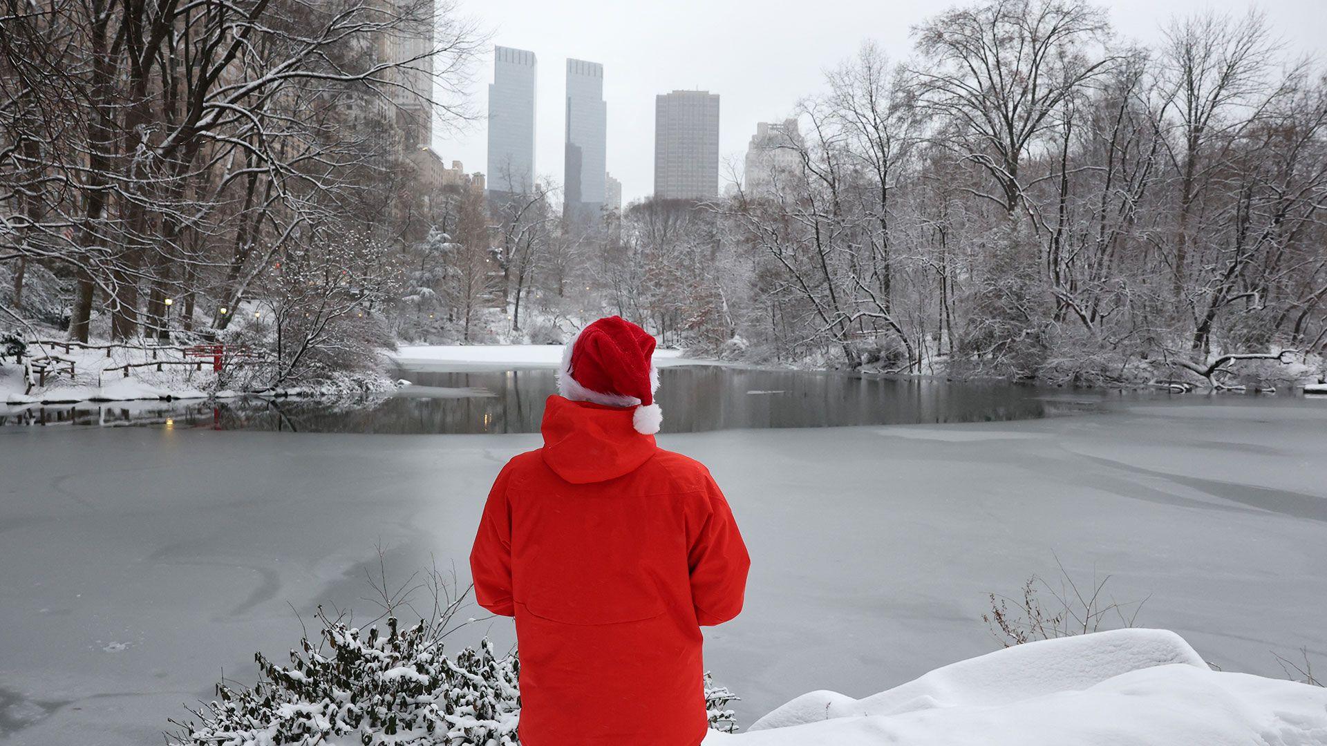 A person in a red coat and Santa hat looks out over an icy lake and snow covered trees with a city skyline in the background