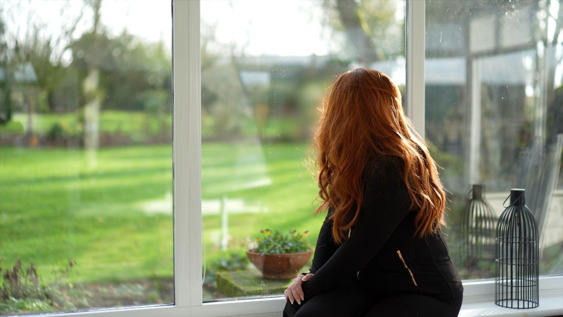 Young woman dressed all in black with bright red hair sits by window with back to reader. 