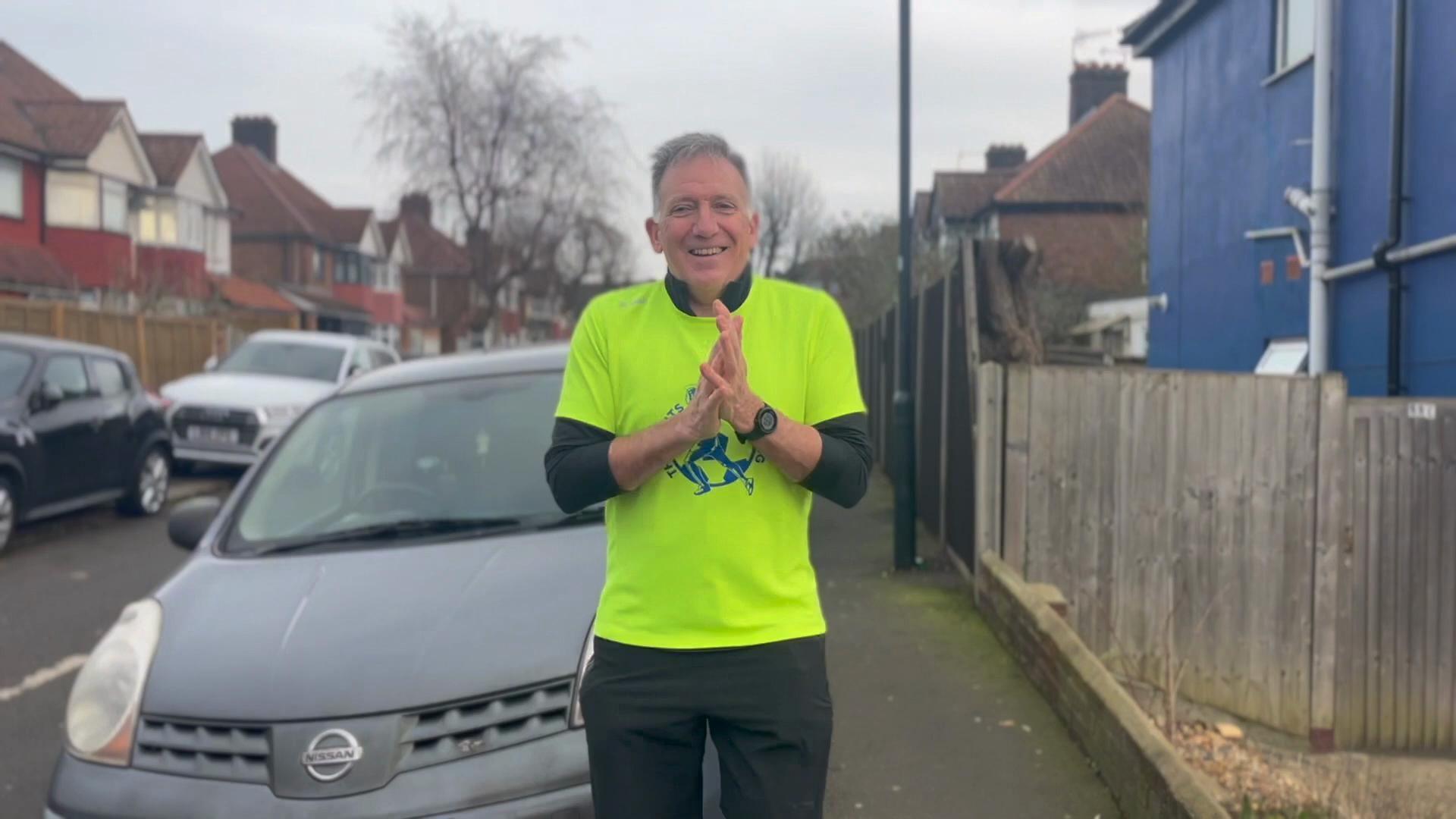 Dave Thomas is standing on a footpath in front of a parked car, wearing his running gear, including a high visibility t-shirt. He has his hands clasped together and is smiling. 