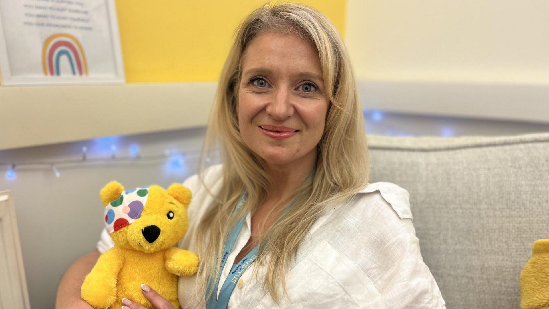 A woman with long blonde hair and blue eyes smiles directly at the camera. She is wearing a white shirt and has a blue lanyard on. She is holding a small Pudsey Bear toy and sat on a cream coloured sofa. In the background - fairy lights and posters can be seen - one has a rainbow on it.