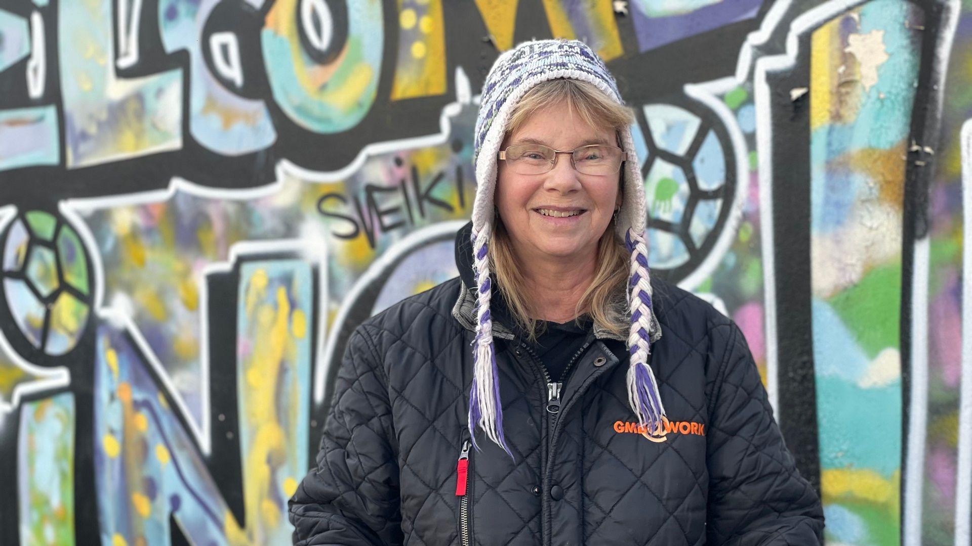 A smiling, blonde haired woman wearing a white and purple patterned hat with wool plaits hanging down onto her shoulders. She wears ovular glasses and a black quilted coat with orange wording and a red tagged zip. Behind her is a multi-coloured graffiti wall with footballs and large lettering.