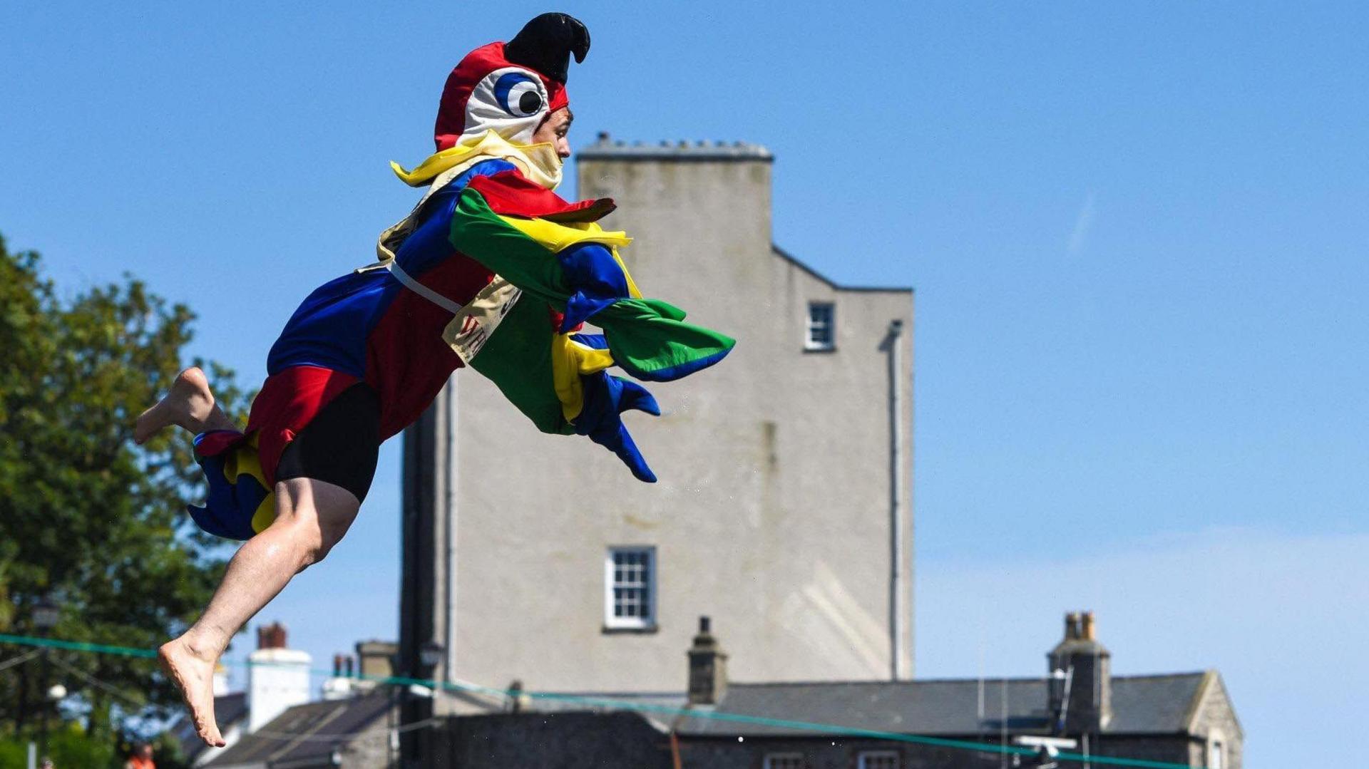A man in a brightly coloured parrot costume appears to float mid-air. He's jumped from the harbourside and is about to land in the sea. The sky is blue and a grey building can be seen in the background.