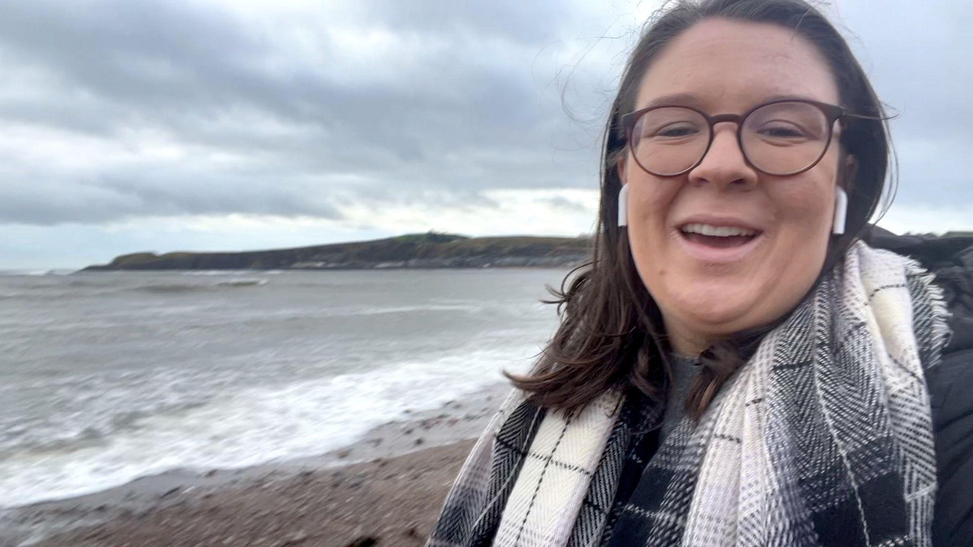 Reporter Catriona Aitken stood with a pebbly beach and sea behind her. She has shoulder-length brown hair and glasses and is wearing a black jacket, black and grey scarf and airpods.