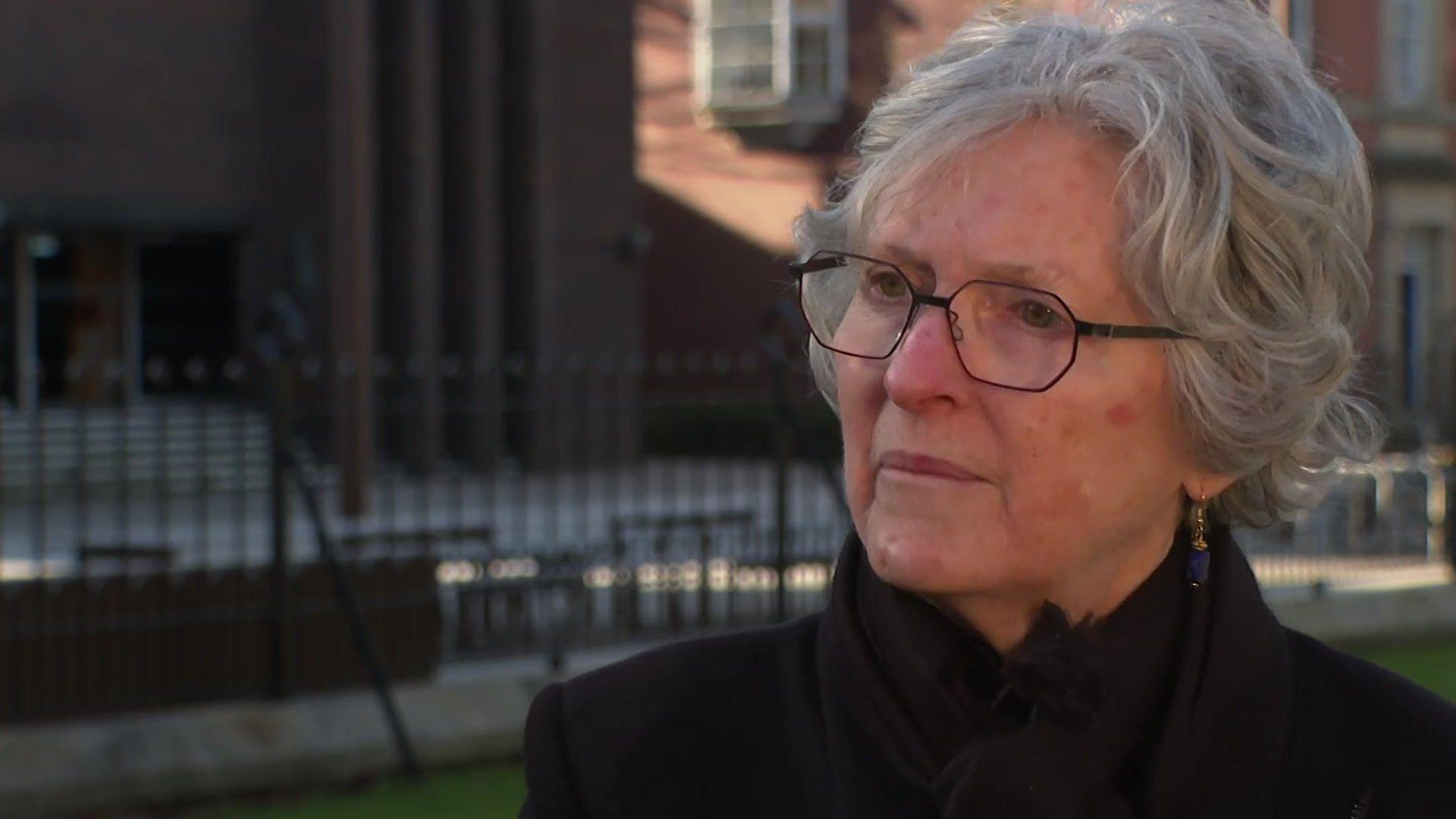 A woman with grey curly hair, and glasses, wears a black coat, and looks to the left of the camera, outside a university building 